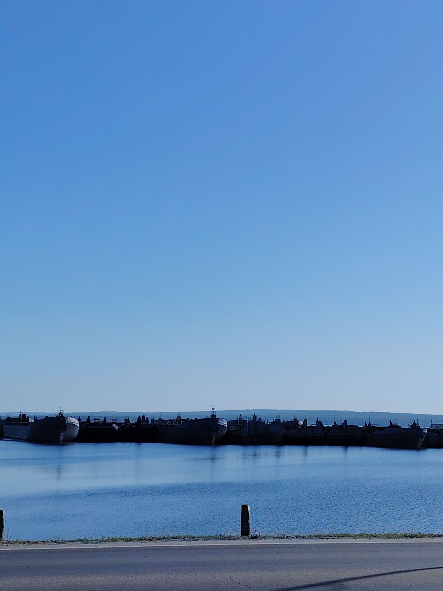 The image depicts a water view with a line of docked ships, likely landing craft, in the city of Chaikovsky, Russia. The water reflects the blue sky. A road with sparse vegetation runs along the shore in the foreground. The absence of distinct details on the ships and the calm water suggest a quiet, still day.