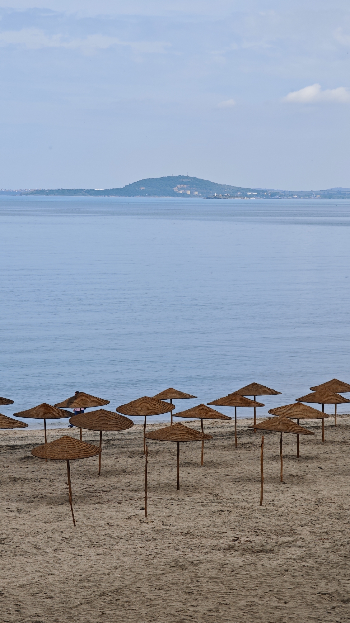 The image shows a beach in Burgas, Bulgaria, with numerous straw umbrellas shading the sand. In the background is the sea, with land visible across the water. The sky is pale blue with a few clouds.
