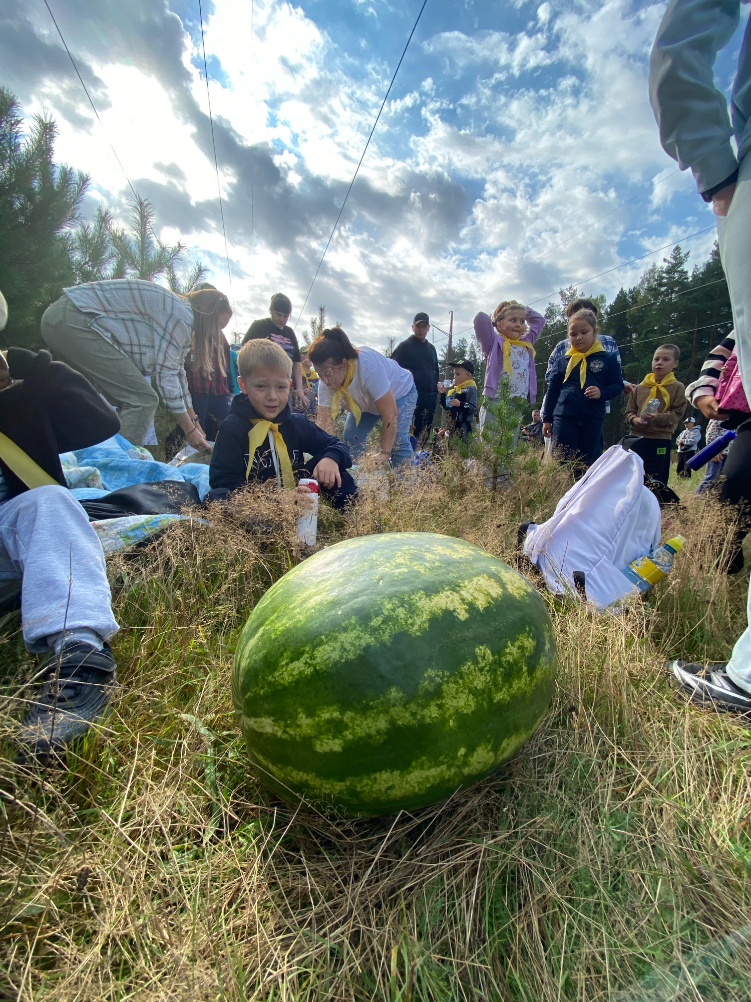 A group of people, including adults and children, are gathered outdoors in a grassy area with dry grass. The sky above is cloudy with patches of sunlight. In the foreground, a large watermelon is prominently displayed. Several children are wearing yellow scarves. One adult is bending down, possibly interacting with a child. Another child is sitting down next to the watermelon. The setting appears to be a casual outdoor gathering or event. The location is identified as Noviy, Russia.