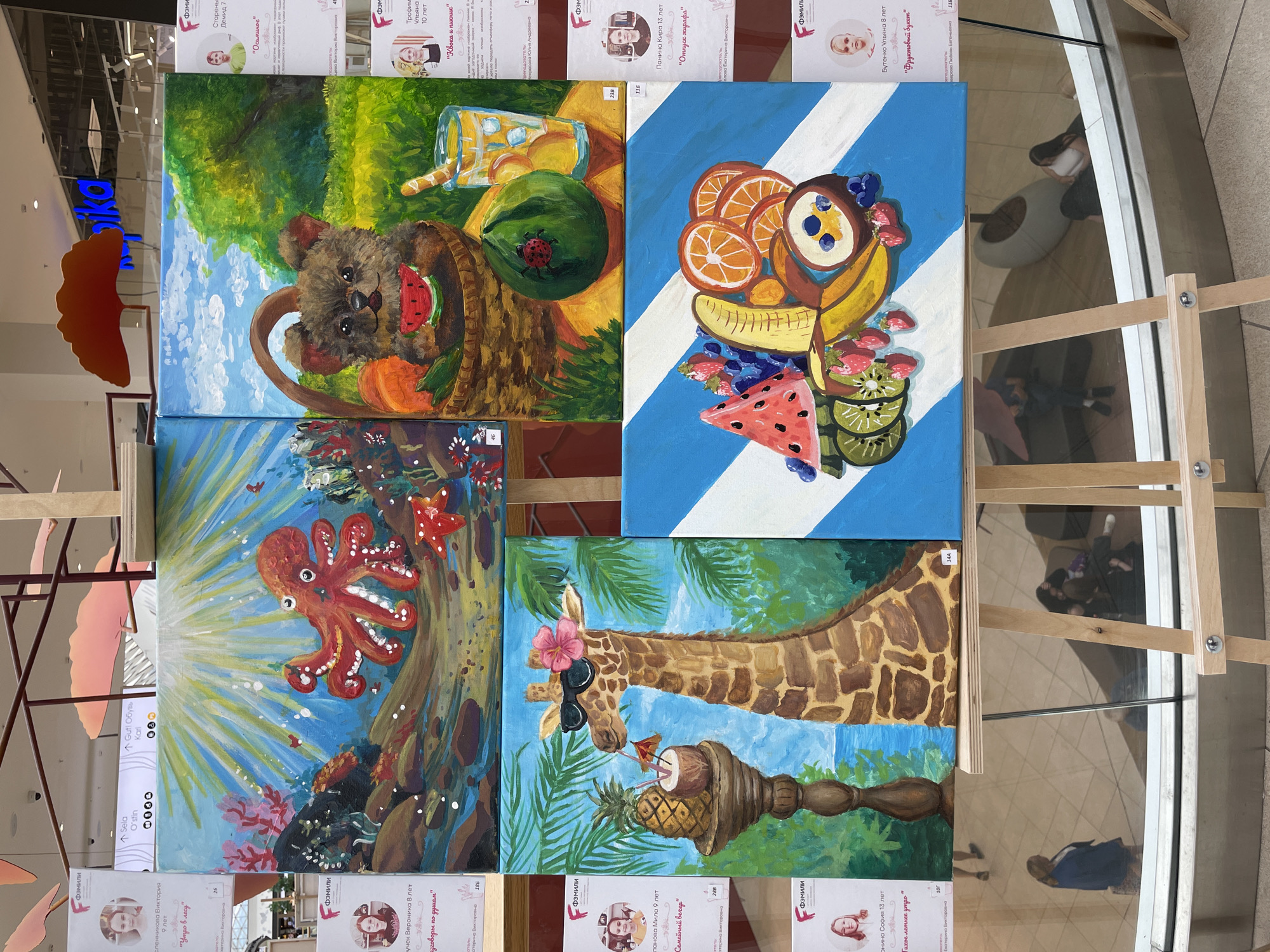 An indoor art exhibition displays several colorful paintings on easels. To the left, a poster board features portraits and titles of artworks, including one by "Skovpich Viktoria, 9 years old" titled "Utro v lesu" (Morning in the forest). Above this, a digital display shows "Sela O'Stin" and "Gut Obyev Kari". In the background, the "Skovpich Viktoria" poster board has a number "26" visible.
The paintings themselves are vibrant and diverse. The top left artwork depicts an underwater scene with a large red octopus. The top right painting shows a teddy bear in a basket eating watermelon, with a glass of lemonade and fruit nearby. The middle left painting features a giraffe wearing sunglasses, holding a coconut drink and pineapple. The bottom painting showcases a collection of fruits, including watermelon, strawberries, oranges, and bananas, arranged on a striped blue and white background.
Several other poster boards with similar formats are visible on the right side of the exhibition, indicating more artworks and their young artists. One of these mentions "Stoyaniv Denis, 7 years old" and "Osinovoe" (Aspen). Another mentions "Troshina Ulyana, 10 years old" and "Klora i pishchye" (Chlora and food). A third mentions "Panina Kira, 13 years old" and "Otpuskaya zhirafo" (Releasing the giraffe). A fourth mentions "Butenko Ulyana, 8 years old" and "Fruktovy duet" (Fruit duet).
The scene appears to be in a public space, possibly a shopping center, given the reflection of people in the background and the overall lighting. The reflection shows individuals browsing the exhibition, with one woman in a blue jacket walking past. The easels are made of light-colored wood.