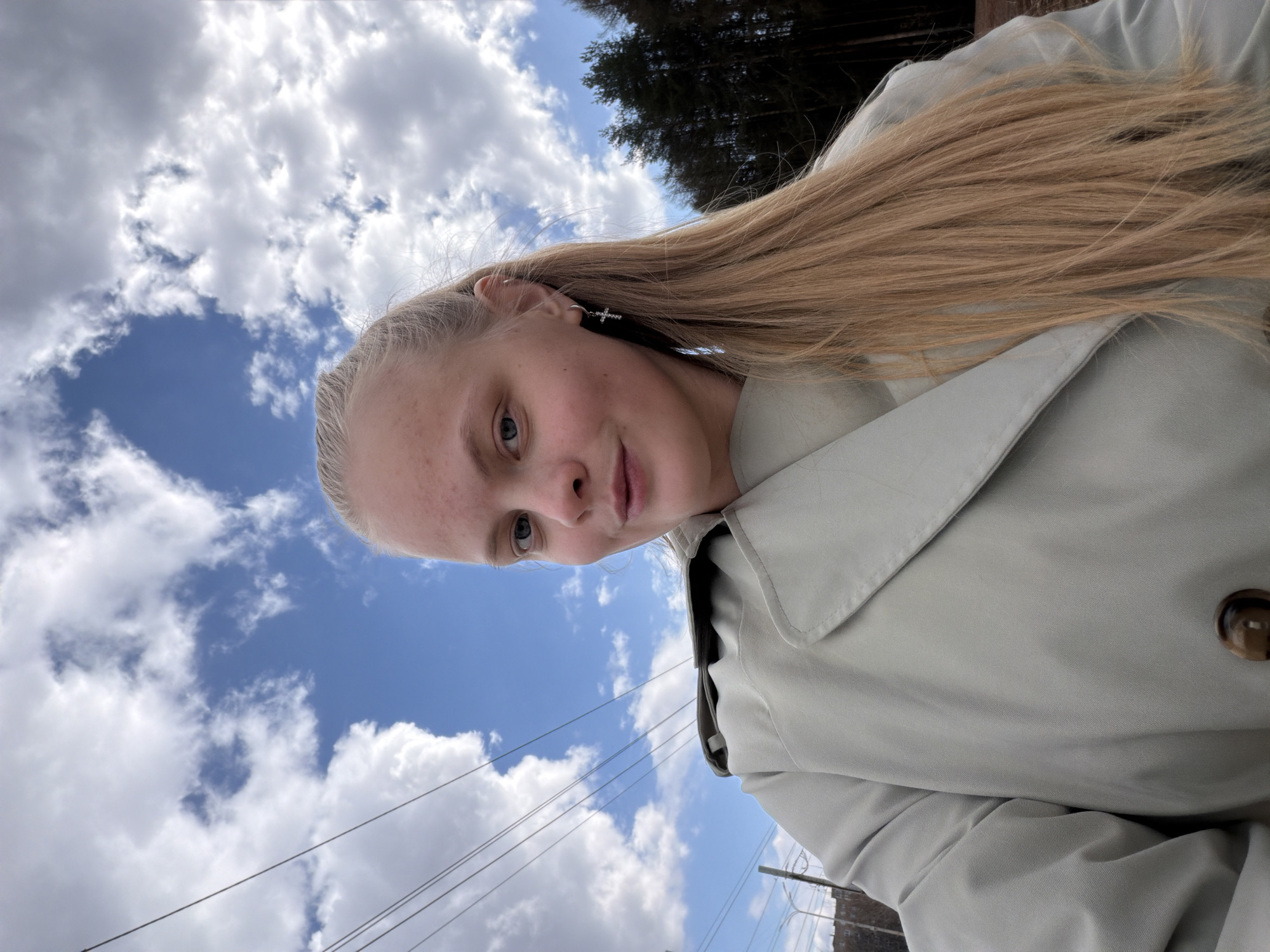 The original image is oriented 90 degrees counter-clockwise. It depicts a young woman in an outdoor selfie taken under a bright, cloudy sky. She has long, straight blonde hair, fair skin with some minor blemishes, and light-colored eyes. She is wearing a light beige or grey trench coat, and her left ear features a small cross-shaped earring and a simple hoop earring. Her expression is calm and neutral, as she looks directly at the camera.
The background predominantly shows a clear blue sky dotted with scattered white cumulus clouds, indicating a bright daytime with fair weather. In the upper right section of the frame, dark green pine trees are visible, suggesting a natural or forested area nearby. In the lower left, utility poles and power lines crisscross the scene, indicating a roadside or semi-urban environment. The image captures a serene outdoor moment. There is no visible text. The provided context indicates the location is Izhevsk, Russia.