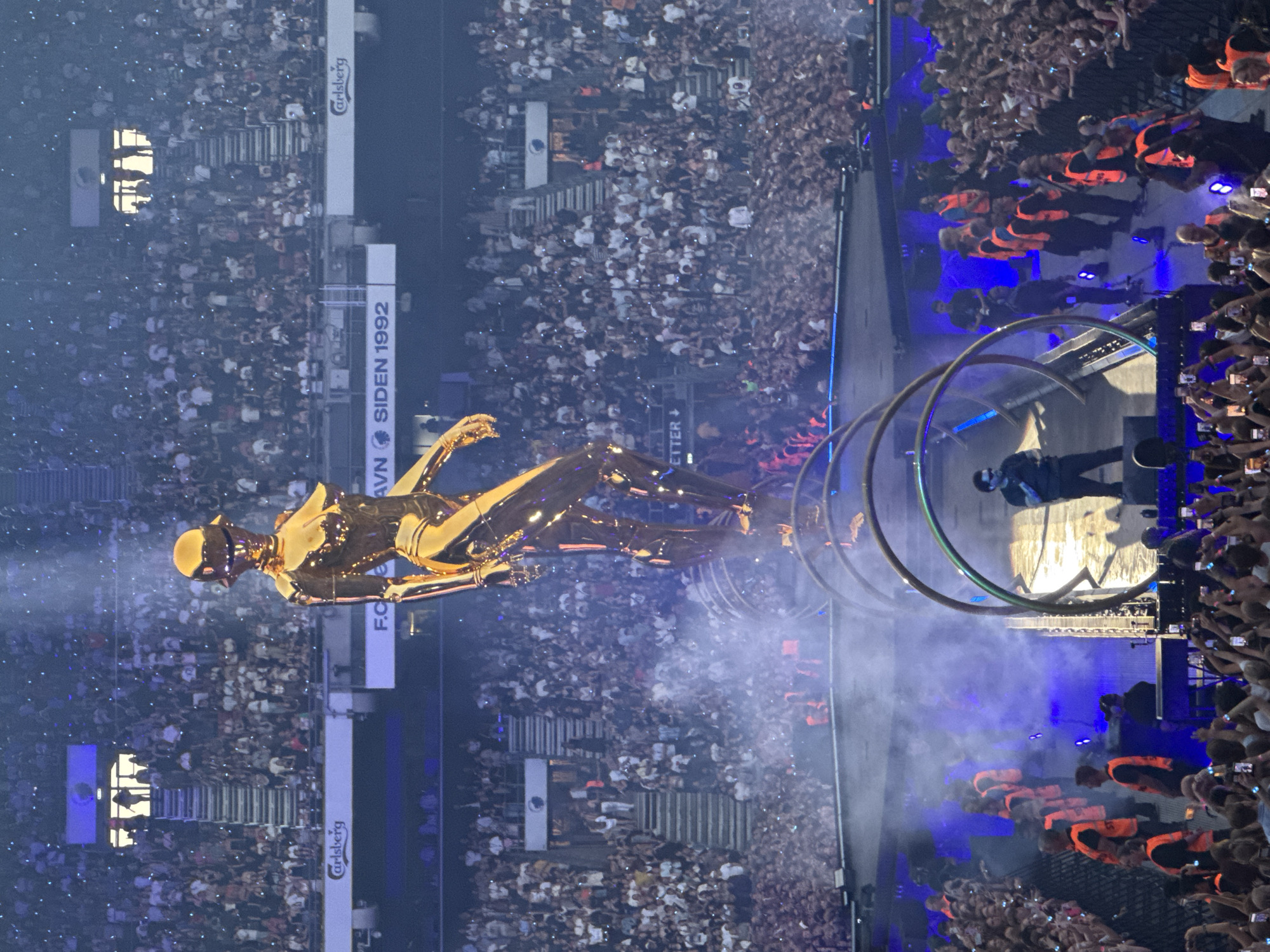 The image captures a large outdoor concert event in Copenhagen, Denmark, likely at Parken Stadium, as indicated by the "F.C. KØBENHAVN SIDEN 1992" signage visible in the background. The central focus is a towering, golden, robotic sculpture positioned on a raised platform. A single performer, dressed in black, stands on a stage below the sculpture, illuminated by spotlights and surrounded by smoke effects. The audience, a vast crowd of thousands, fills the stadium seating and the area around the stage, with many individuals holding up mobile phones. Security personnel, identifiable by their orange vests, are visible at the perimeter of the stage area. The overall scene suggests a vibrant and energetic live performance.