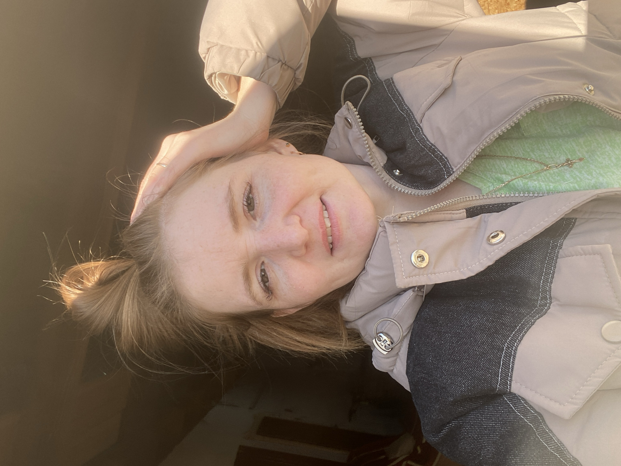 A woman with fair skin and reddish-blonde hair is taking a selfie. She is wearing a light brown padded jacket with dark denim accents, under which a light green top and a gold necklace are visible. Her hair is partially tied up in a small bun. She is holding her right hand on her head. The background is dark and indistinct, with some blurry hints of wooden structures. The location is assumed to be in the city of Chaikovsky, Russia.