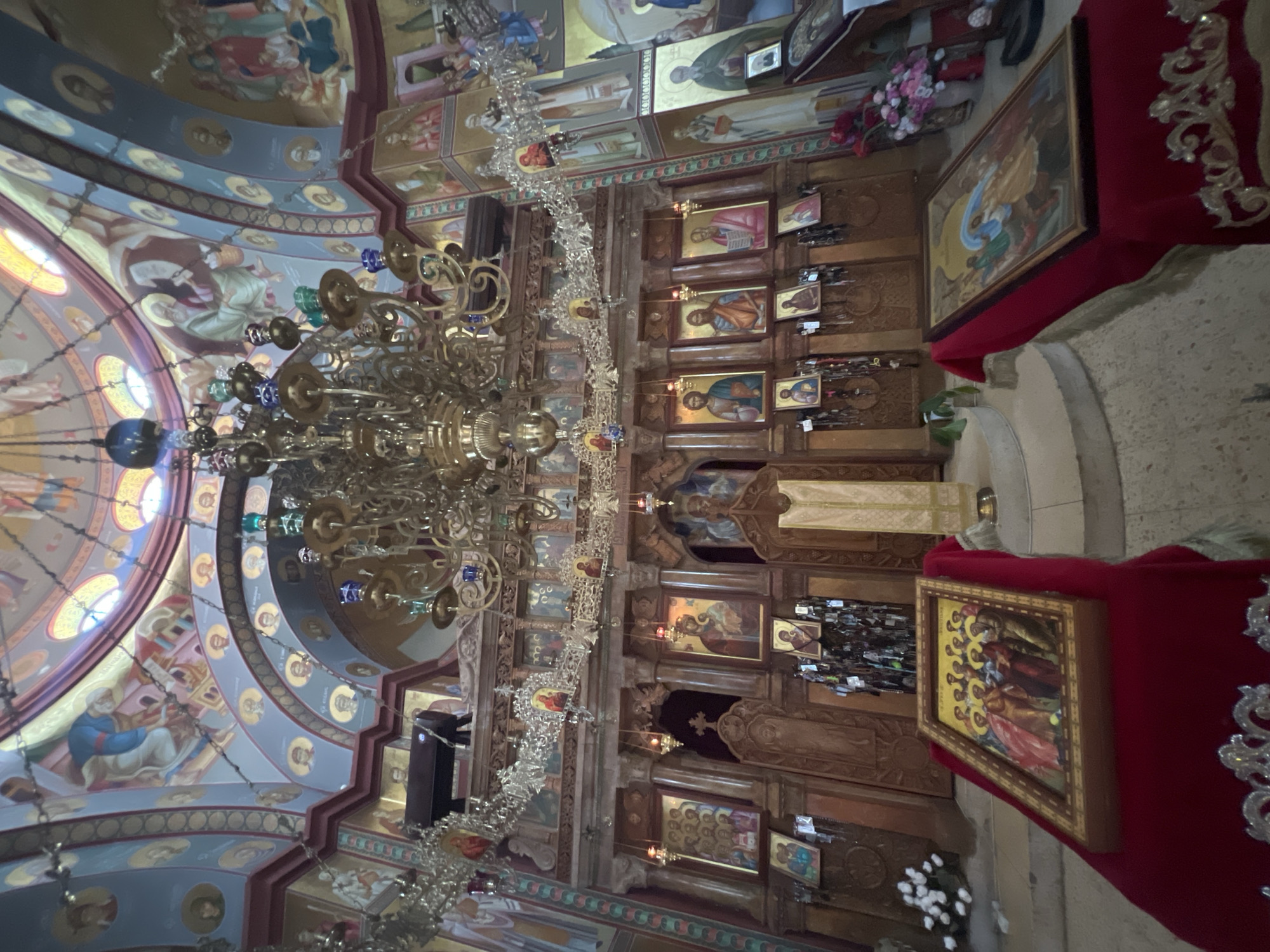 This is an interior view of a richly decorated Orthodox Christian church, likely in Emek HaYarden Regional Council, Israel, given the context. The scene is dominated by an ornate iconostasis, a wooden screen covered with religious icons, that separates the nave from the sanctuary. Above the iconostasis hangs a large, multi-tiered brass chandelier with colorful glass accents. The walls and domed ceiling are adorned with vibrant religious frescoes depicting biblical scenes and figures.
In the foreground, two large framed icons rest on red velvet-covered stands. The left icon depicts a group of figures, possibly apostles, while the right icon shows a winged angelic figure. Behind the iconostasis, a clergyman in white and gold vestments stands on a raised platform. Various smaller icons are placed throughout the scene, along with decorative elements like carved wood and floral arrangements. The floor appears to be made of stone or tile. The lighting is subdued, with light filtering in from the dome and from lamps within the church.