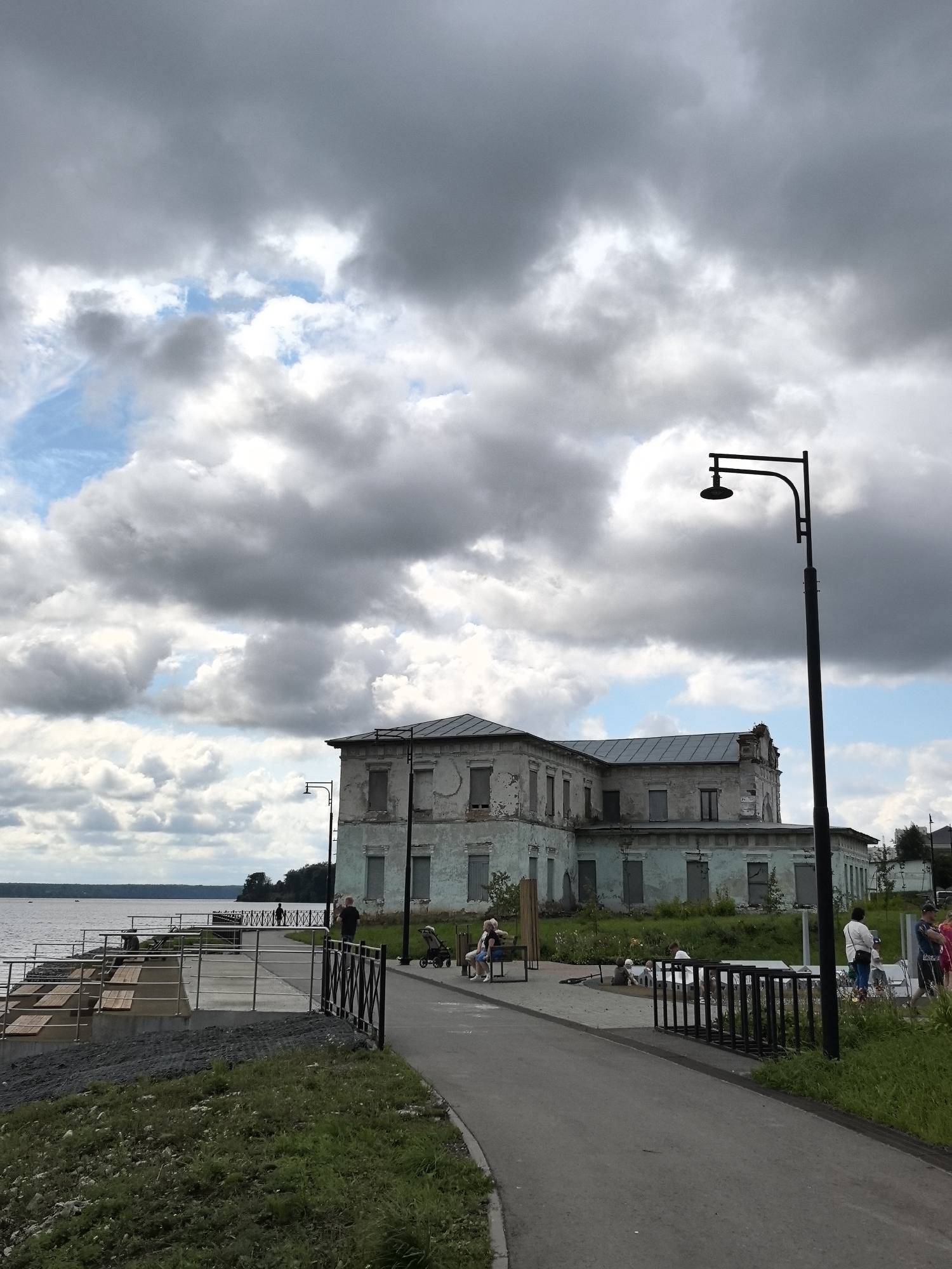 An outdoor, low-angle, medium shot captures a cloudy day at the embankment in Dobryanka, Russia. A weathered, two-story building with boarded-up windows stands prominently on the right, its pale blue facade peeling and showing signs of disrepair. A tall, black lamppost with a curved arm stands in front of the building. A paved walkway curves into the foreground, leading towards the building and a waterfront promenade.
The promenade, on the left, features metal railings and wooden benches. Several people are visible along the promenade and walkway. On the promenade, two figures stand near the water's edge, and further along, a solitary silhouette is seen. Closer to the building, a woman is seated on a bench with a child. On the walkway, a man pushes a stroller, and a group of people, including a woman in a white shirt and a man in a blue shirt, are gathered. A child sits on the ground near a small bench. In the background, a body of water, likely the Kama River, stretches out under a dramatic sky filled with dark, heavy clouds interspersed with patches of bright blue. Green grass and some vegetation grow along the embankment and around the building.