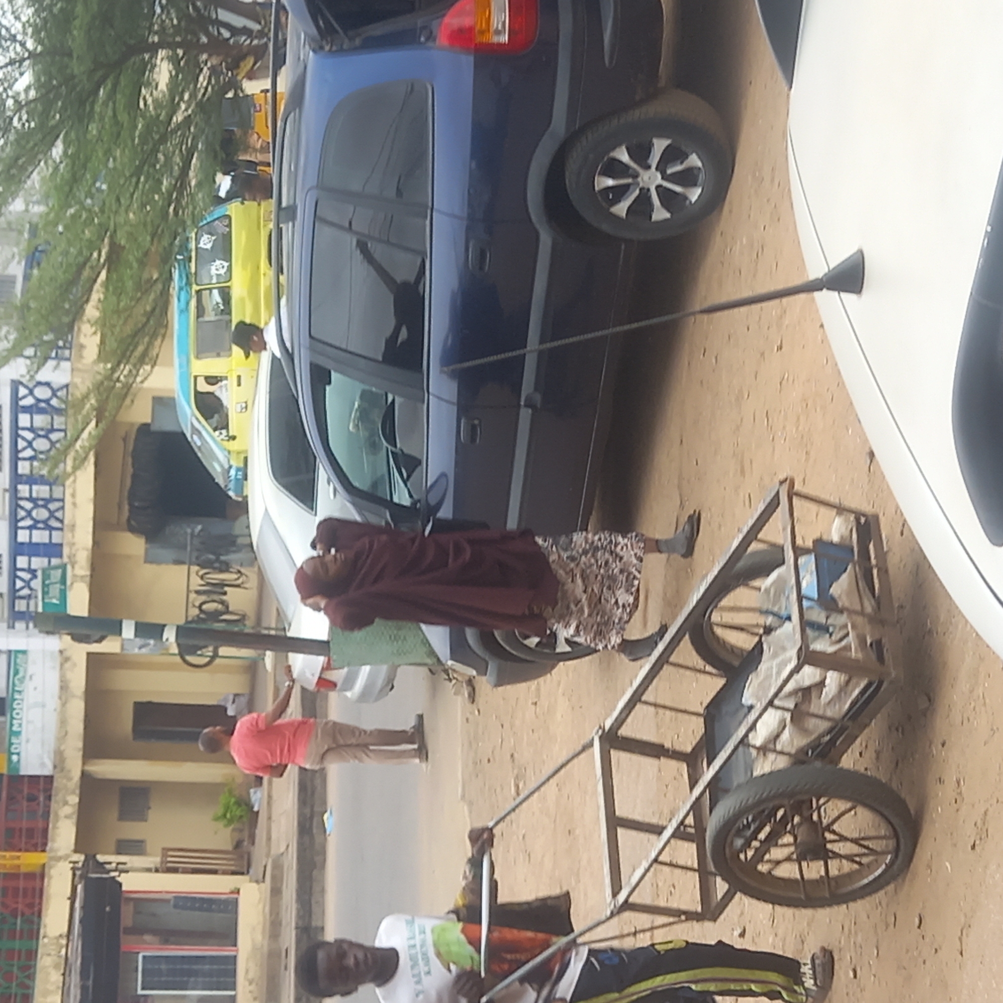 The image depicts a street scene in Dadi Riba, Nigeria. Several vehicles, including a blue van and a white car, are parked along the street. A yellow and blue public transportation vehicle is visible in the background. A woman in a burgundy robe and floral skirt stands near the blue van. A man in a pink shirt is standing on the sidewalk. Another man is pulling a metal cart with two wheels. Buildings are visible in the background, including a structure with blue and white patterned details. The ground appears to be a sandy or dirt surface.