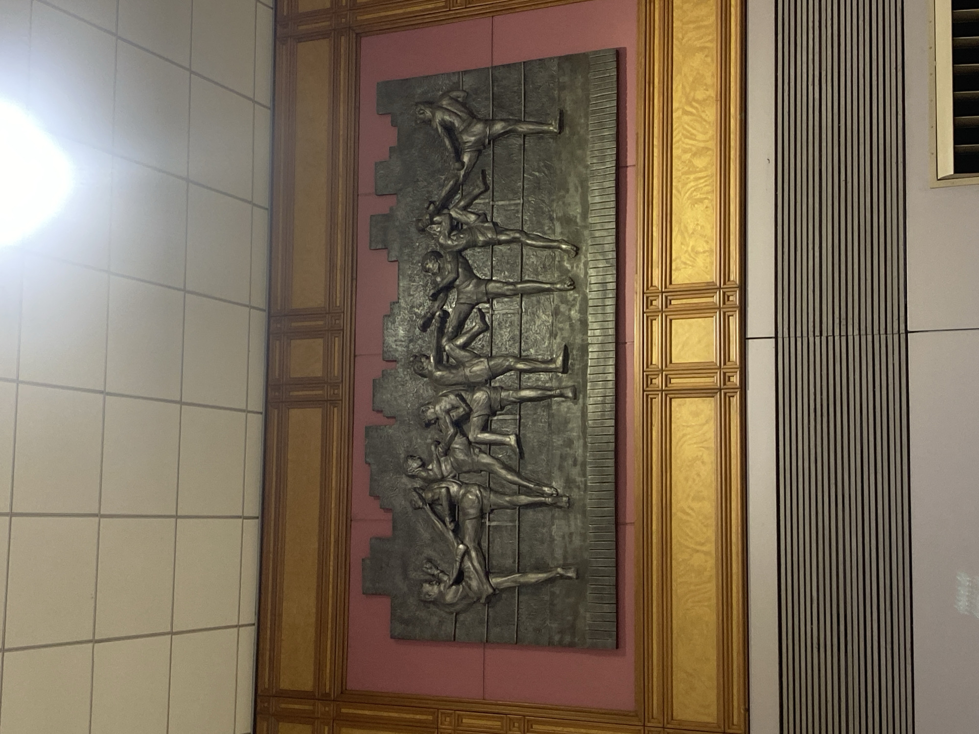 An interior shot captures a large metallic bas-relief sculpture mounted on a wall, depicting several human figures in dynamic, athletic poses. The sculpture is oriented vertically, showing figures seemingly climbing or struggling upwards, with bodies overlapping and limbs extended. There are approximately seven or eight distinct figures visible in the relief, each rendered with strong musculature and an impression of effort.
The sculpture is set against a flat, reddish-pink wall panel. This panel is framed by ornate golden decorative elements, featuring intricate patterns and mouldings that run horizontally and vertically, creating a grand, structured border around the artwork. To the left of this framed section, a brightly lit wall covered in light-colored square tiles is visible. To the right, there is a section of wall adorned with closely spaced vertical dark slats, suggesting a vent or a textured architectural feature. A small part of a louvered vent is also visible in the upper right corner.
The lighting in the scene is artificial, with a bright light source creating a strong glare on the tiled wall to the far left. The overall setting appears to be a public interior, possibly a station or institutional building, characterized by robust materials and decorative art. No visible text is present in the image. The context provided indicates the location is Bangkok, Thailand.