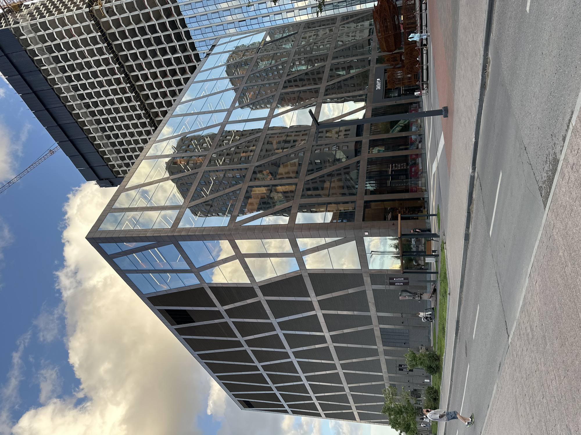 This is a photograph of a modern, angular building with a dark facade and a distinctive pattern of glass windows on its exterior. The building is reflecting a multi-story residential building with a honeycomb-like exterior. In the background, a tall construction crane is visible, indicating ongoing development in the area. The sky above is partly cloudy with a blue sky visible. In the foreground, a street with a pedestrian crossing is present, and a man is walking across the street. The building appears to be a commercial or office space, possibly part of a larger development in a city. The signage on the building includes "МУЗЕИ" (MUSEUMS) and "ЗИЛАРТ" (ZILART), suggesting the location is the ZILART district in Moscow, Russia, which is a developing residential and cultural area on the former grounds of the Likhachev automobile plant.