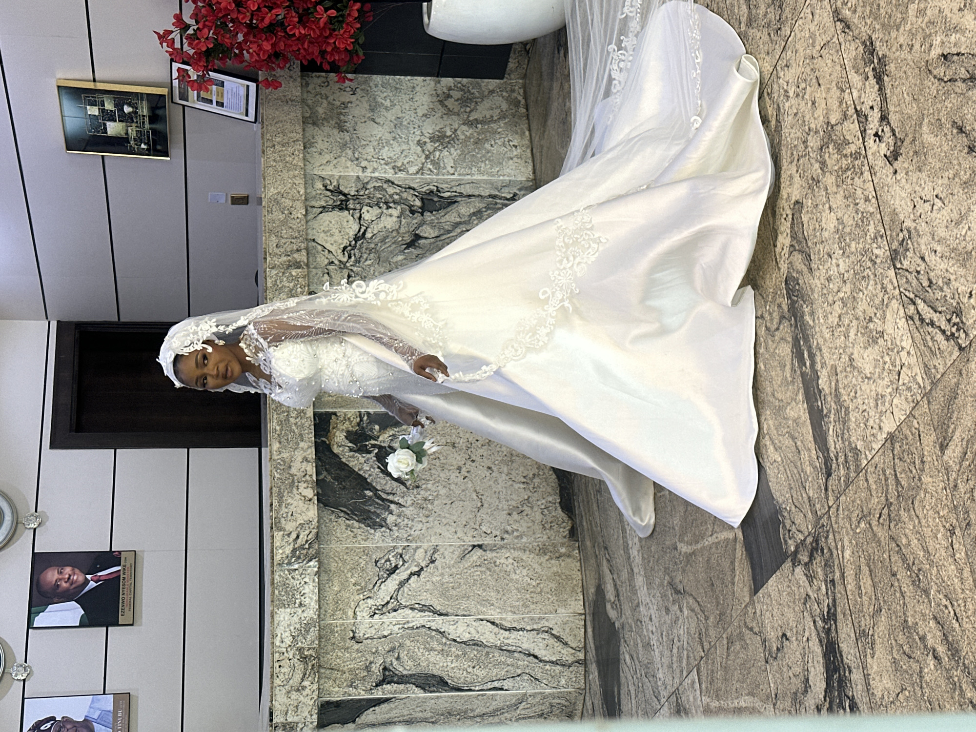 A bride in a white wedding dress stands in what appears to be a public building lobby, possibly in Abuja, Nigeria. The dress features a long train, lace detailing on the sleeves and bodice, and a veil. She is holding a small bouquet of white roses. Behind her, there is a marble counter with framed pictures on the wall above, one of which appears to be a portrait of a man in a suit and tie. To the right, a tall white vase holds a vibrant arrangement of red artificial flowers. The floor is tiled with a granite-like pattern. The overall impression is of a formal setting, likely a reception area or government building given the portraits.