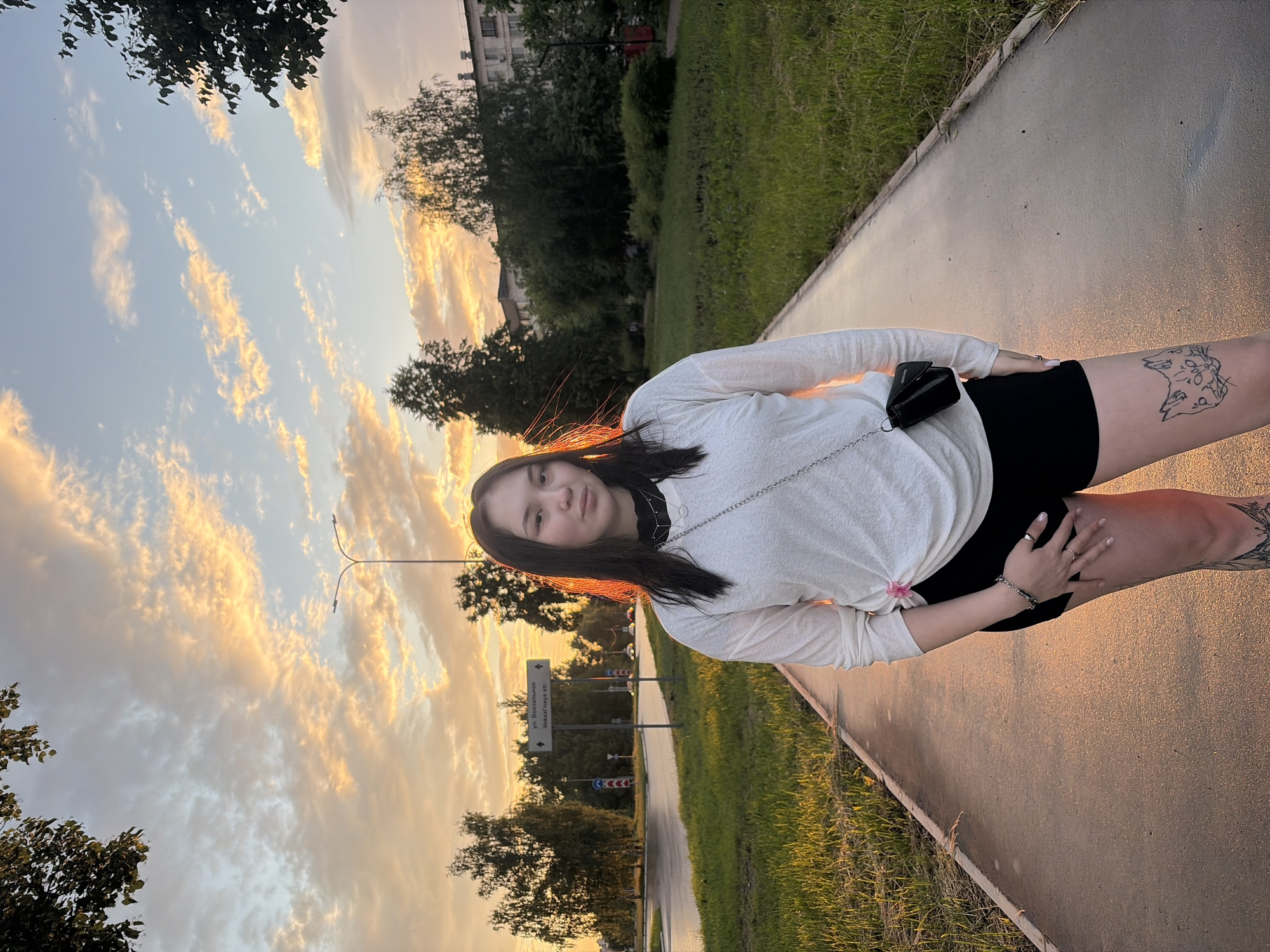 A young woman stands on a paved pathway at sunset in Chaikovsky, Russia. She has long dark hair and is wearing a white long-sleeved shirt, black shorts, and a small black purse across her body. She has tattoos on her left thigh, one depicting two cats. The pathway is bordered by green grass and trees. In the background, there is a street with traffic, buildings, and a street lamp. The sky is filled with dramatic clouds illuminated by the setting sun, casting a warm glow over the scene. A street sign with Russian text is visible to the left of the woman.