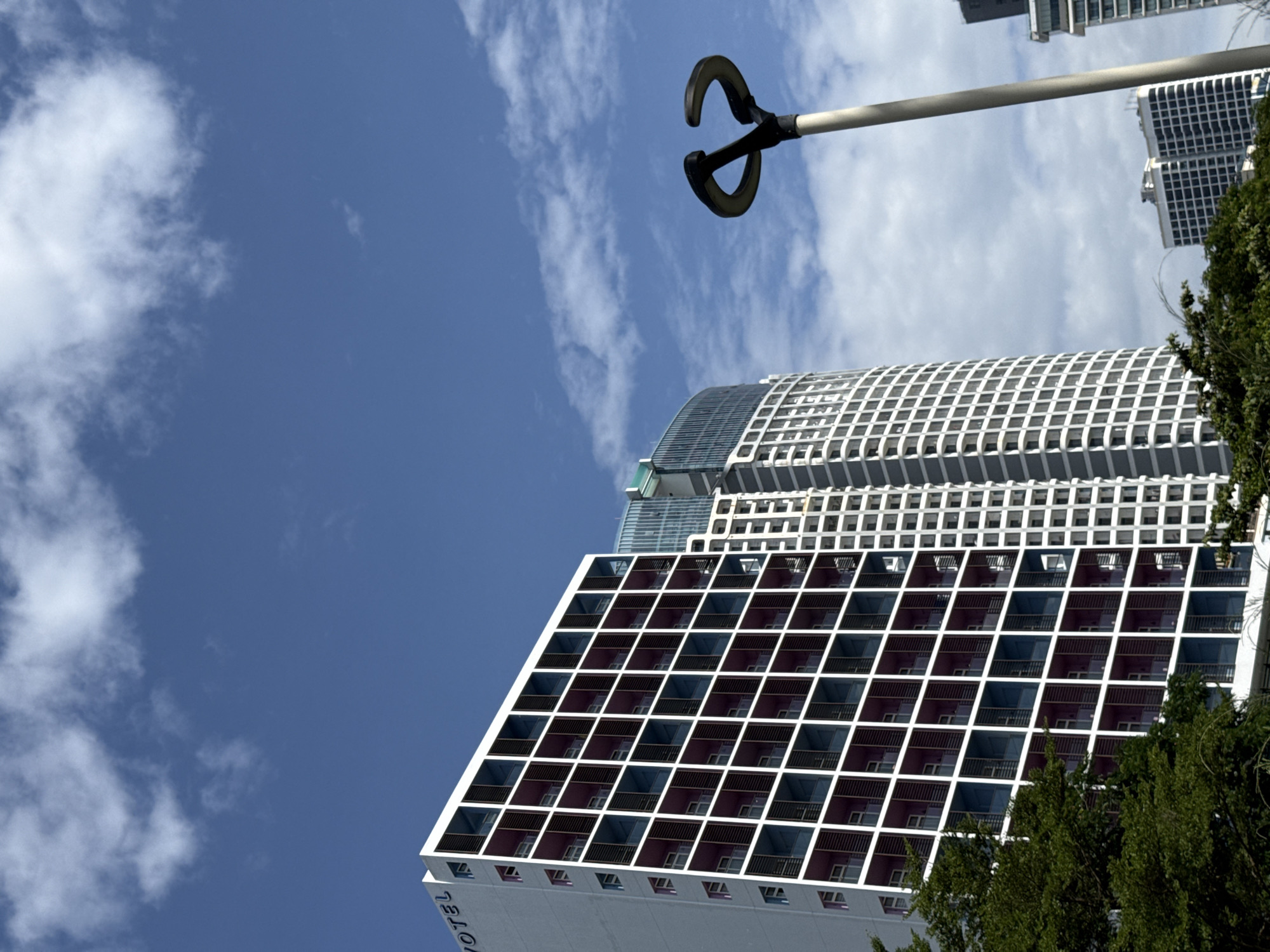 A low-angle, vertical medium shot captures a modern hotel and surrounding buildings against a bright blue sky with scattered white clouds. The prominent building on the left is the Novotel Nha Trang hotel, a tall, rectangular structure with a distinctive facade. Its exterior is white, punctuated by a grid of square balconies. Each balcony has white railings, and the interiors are colored in alternating shades of deep purple and dark blue. The "NOVOTEL" logo is visible in blue lettering on the upper left side of the hotel's facade.
To the right of the Novotel, a taller, more rounded skyscraper with a glass and white facade rises, its upper sections obscured by the sky and clouds. Further in the distance, more buildings of varying heights and architectural styles are visible, including several white, multi-story structures with repetitive window patterns. In the foreground, the tops of lush green trees frame the lower left corner of the image. A modern street lamp with a distinctive curved design stands tall on the right side, its pole a light gray and the curved lamp heads a dark color, silhouetted against the sky. The overall impression is one of urban development and a clear, sunny day in Nha Trang.
