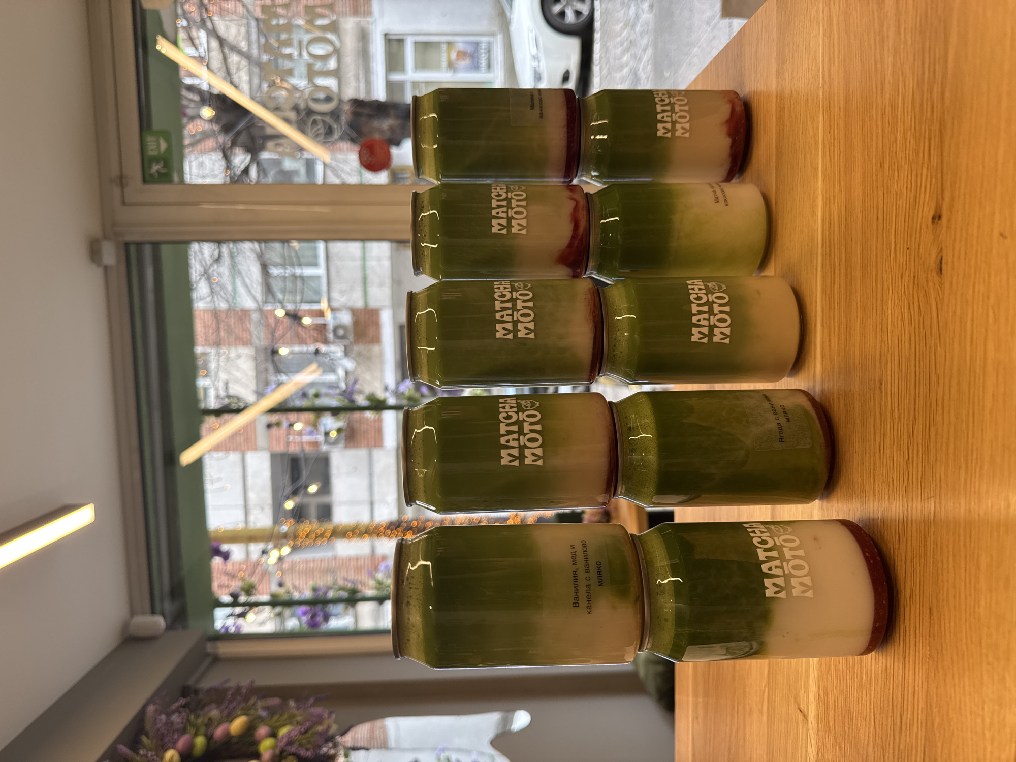 The image shows several cans of Matcha Moto drinks lined up on a wooden surface. The cans are stacked in pairs, with each pair consisting of two different flavors. The flavors are "Vanilla, Honey, and Cinnamon with Vanilla Milk", "Strawberry with Vanilla Milk", "Raspberry Matcha Coconut", and "Matcha Moto" written in white capital letters with a logo including a leaf on some green, glass-like cans. The background features a window with a view of a street scene, including buildings and parked cars. The location is identifiable as Sofia, Bulgaria, due to the architectural style of the buildings. There are additional decorations in the window, including flowers and lights. The environment is bright and indoor.