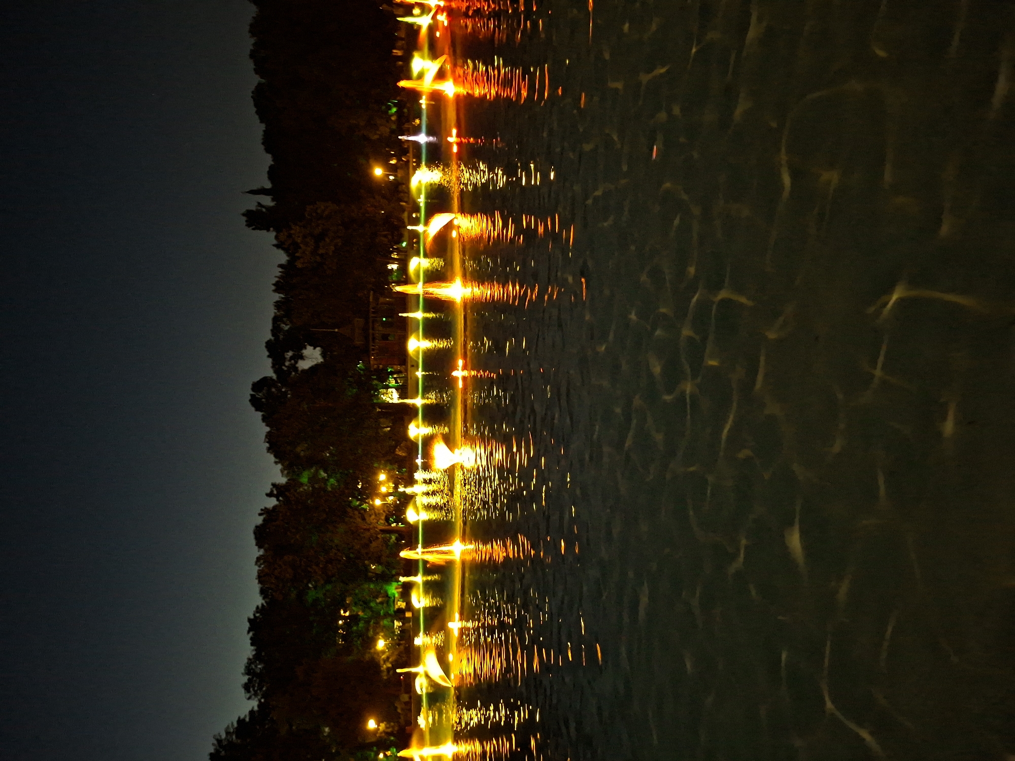 The image depicts a night scene of the Singing Fountains in the city of Plovdiv, Bulgaria. The fountains are illuminated with colorful lights, creating a vibrant spectacle against the dark sky. The water from the fountains is captured in motion, with streams of water reaching upwards and outwards, reflecting the colorful lights onto the surface of the pool. Trees and lights from the surrounding area are visible in the background, silhouetted against the night sky. The overall impression is one of a lively and beautiful urban landscape at night.