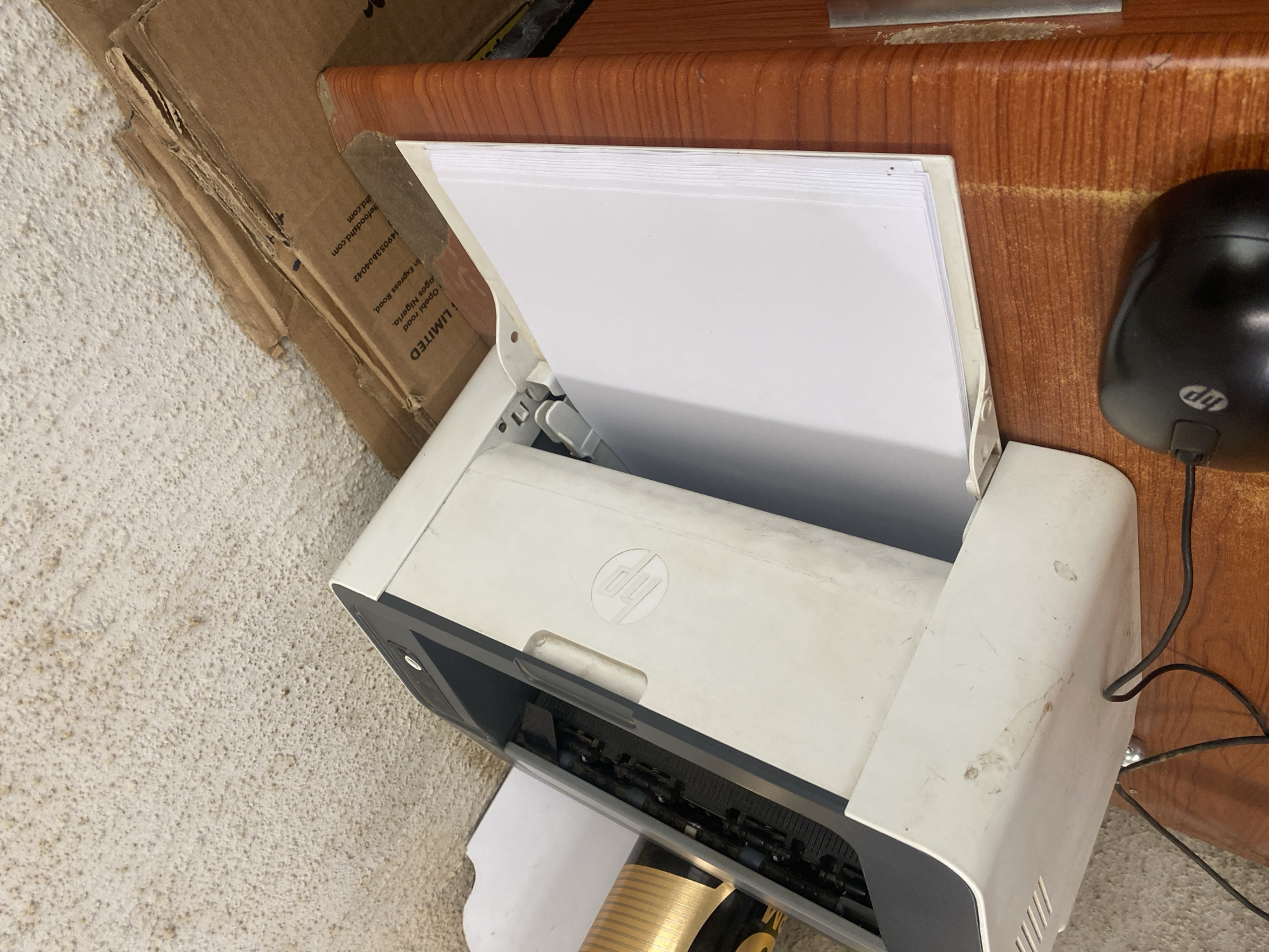 The image shows a white HP printer with a stack of white paper in its input tray. The printer is open, revealing its internal mechanisms. A black HP computer mouse is visible to the right of the printer, placed on a wooden surface. To the left of the printer, a torn cardboard box is leaning against a textured, light-colored wall. Text is visible on the cardboard box, including "LIMITED", "Opelba road", "Iagos Nigeria", and a website address "herfoodbird.com" along with a phone number. The scene appears to be indoors, possibly in an office or home environment. The lighting suggests it is daytime. The overall impression is that of a workspace with an active printer and surrounding materials.