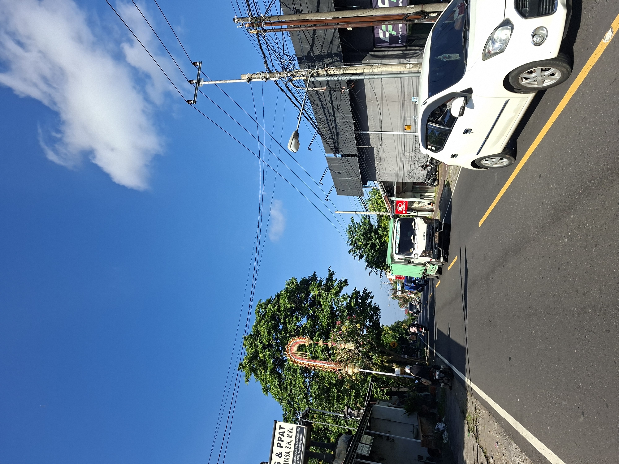 This image depicts a street scene in Banjarangkan, Indonesia, under a clear blue sky with scattered white clouds. The road is asphalt, with a yellow line marking the center lane and a white line bordering the right side. On the left side of the road, there is a sidewalk and buildings, including one with a sign that reads "S & PPAT MAYASA, S.H., M.Kn.". A large green tree stands prominently behind a decorative archway.
Several vehicles are present: a white car is in the foreground on the right, a green Isuzu truck is in the middle of the road, and several motorcycles are visible further down the street. People are seen riding on the motorcycles. Electrical poles with numerous wires crisscross the sky above the buildings. The overall environment suggests a busy urban or semi-urban area.