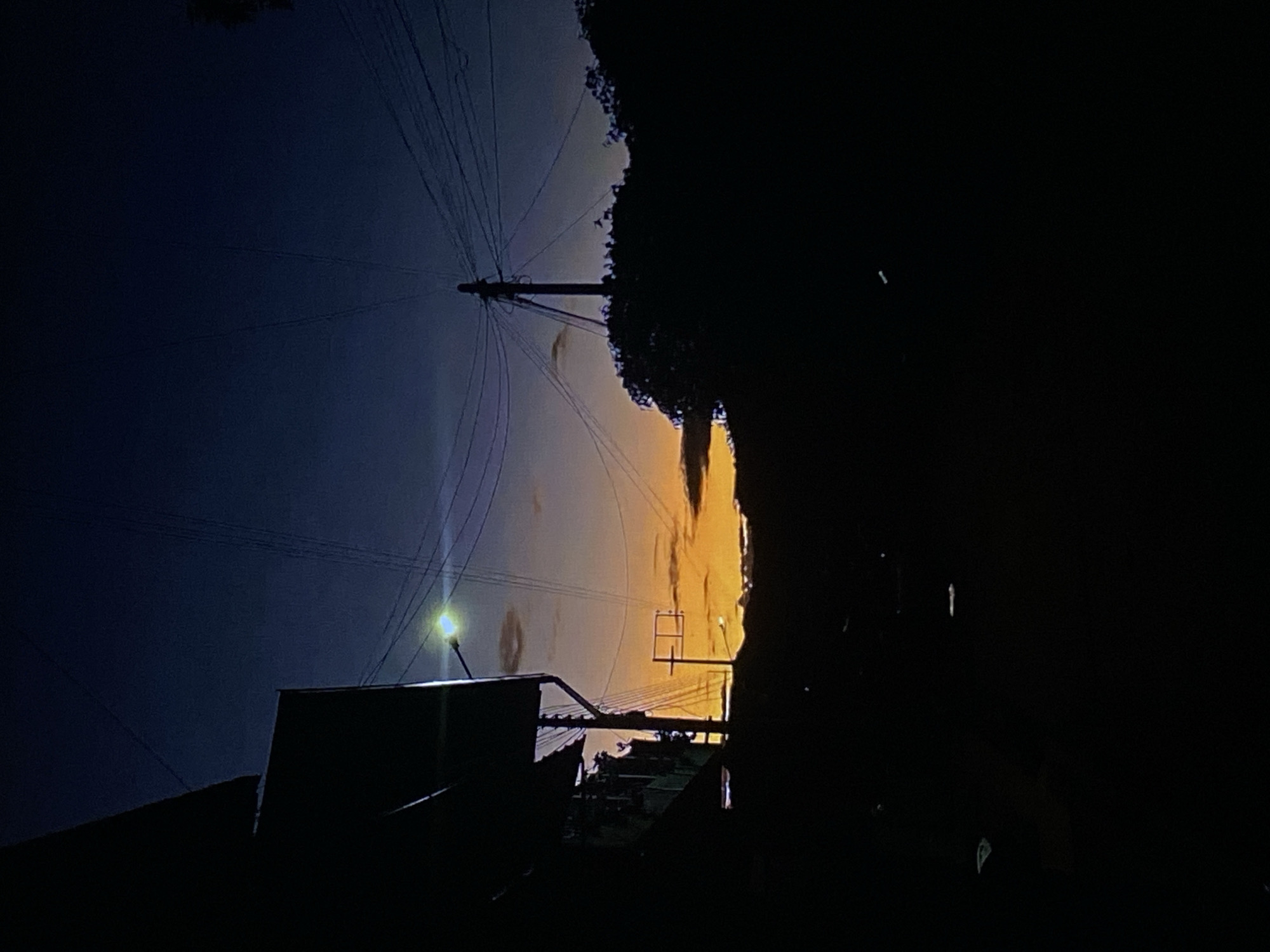 The image captures a twilight scene, likely dusk, characterized by a striking color gradient in the sky. The left side of the frame features a deep indigo blue sky, which transitions horizontally to a vibrant orange and yellow glow on the right. A prominent, dark utility pole is silhouetted near the center of the image, from which numerous power lines extend across the blue sky to the left and into the darker regions on the right.
In the lower-left quadrant, a bright streetlight emits a distinct, slightly greenish-yellow light, partially illuminating a dark, rectangular structure below it. Another, smaller light source is visible further to the right, amidst the orange sky. Dense, dark foliage or trees are silhouetted along the right edge of the image, blending into a large, dark, undefined mass that occupies the entire rightmost portion of the frame and the bottom. Below the main utility pole and power lines, the dark silhouettes of other buildings or rooftops are faintly visible against the lighter sky.
The static scene depicts an urban or semi-urban environment, with no discernible human activity. The active streetlights and the dramatic sky gradient strongly indicate either dawn or dusk, with dusk being more probable due to the streetlights being on. There is no visible text. The overall atmosphere conveyed is calm and contemplative, capturing the transition from day to night in Tinaquillo, Venezuela.