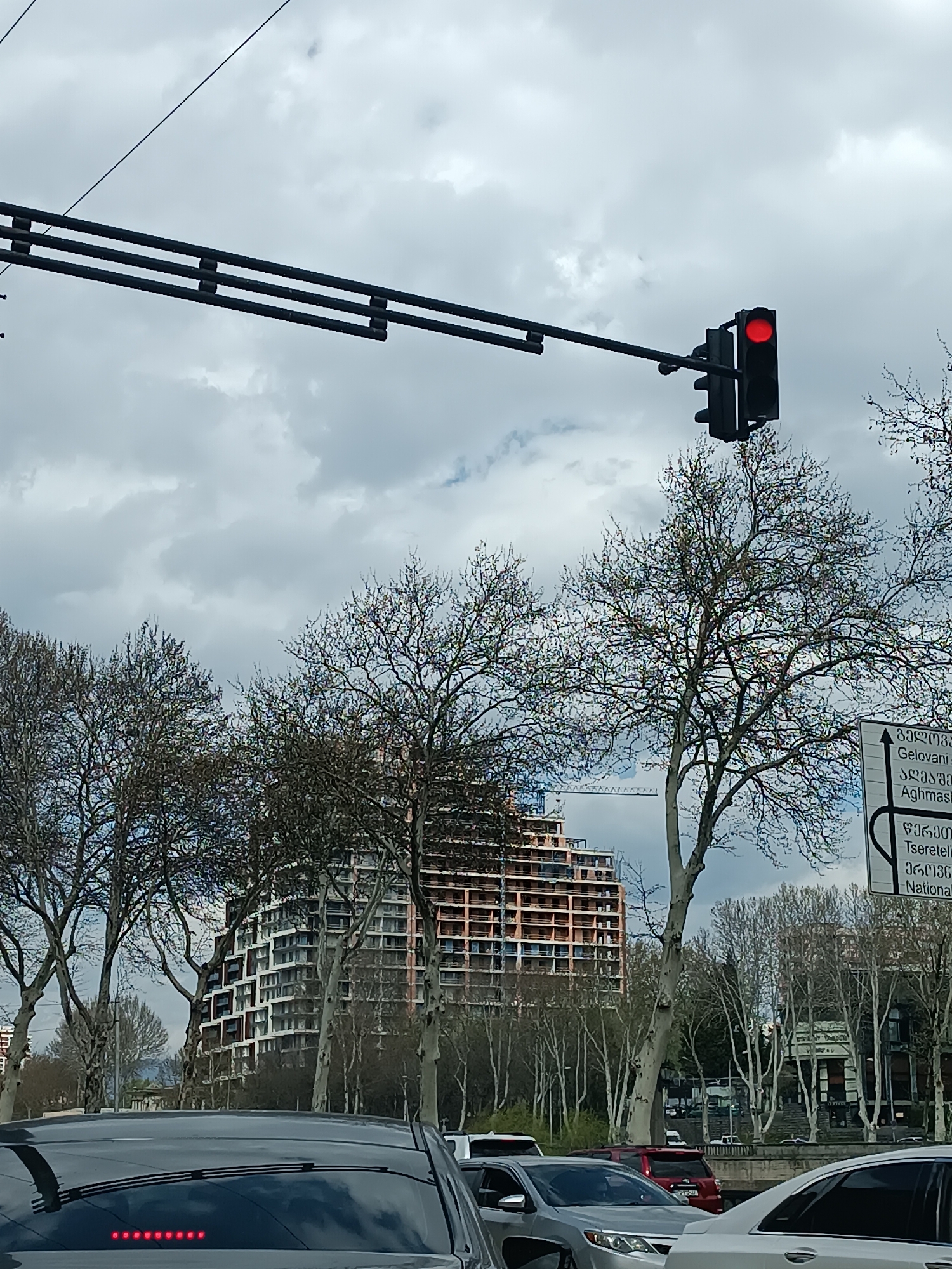 The image depicts a street scene in Tbilisi, Georgia, characterized by a red traffic light, cars in traffic, trees, and a building under construction. The sky is cloudy. A signpost with Georgian text is also visible. The prominent building under construction is a large structure with a terraced design. The image suggests a bustling urban environment with ongoing construction activity.