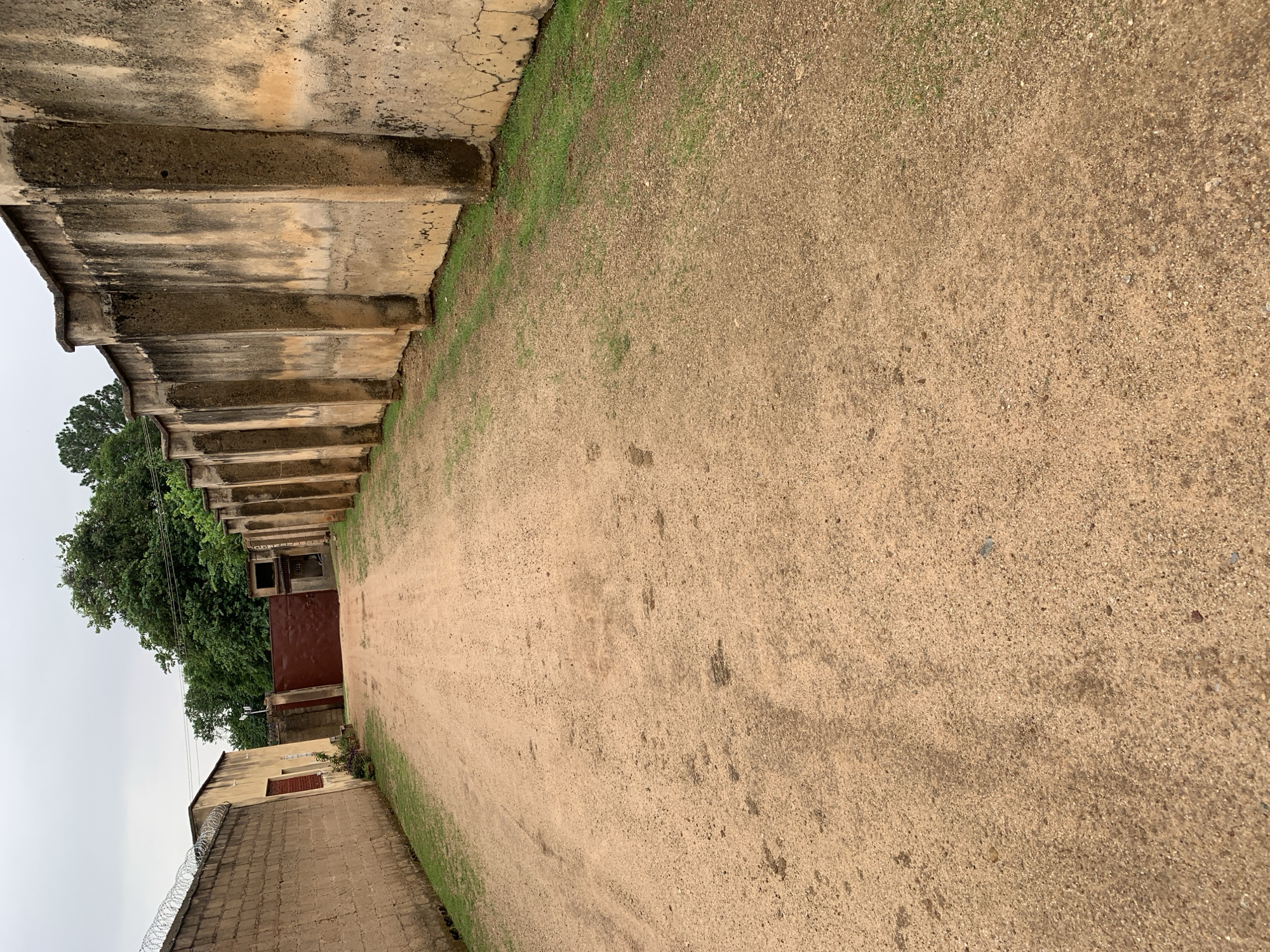 A dirt road in the city of Jos, Nigeria, leads toward a large, closed, dark red gate. On the left side of the road, a tan brick wall topped with barbed wire runs along the edge of a building. On the right side of the road, a tall, aged concrete wall with a weathered, mottled texture extends into the distance. Patches of grass grow along the base of both walls. The sky is overcast.