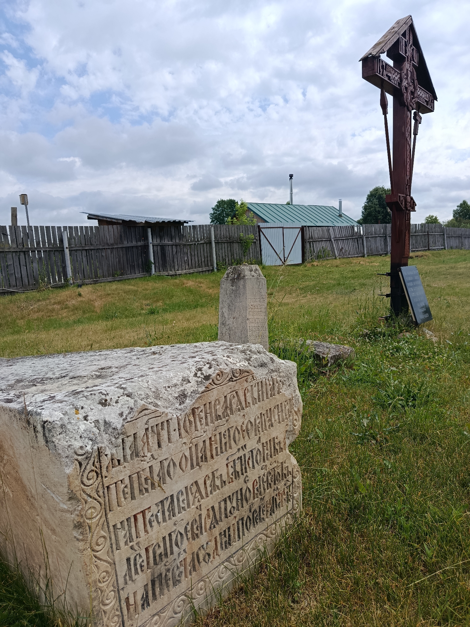 The image depicts a cemetery located in Radovitsy, Russia. In the foreground, a large, weathered stone slab with intricate carvings and Cyrillic inscriptions lies on the grass. To the right, a tall wooden cross stands prominently, adorned with religious imagery and more inscriptions. Behind these markers, a rustic wooden fence encloses the cemetery grounds. Beyond the fence, a simple building with a green metal roof and a chimney suggests a rural or historical setting. The sky above is partly cloudy, casting an overcast light on the scene. The environment is characterized by overgrown grass and a sense of age and history.