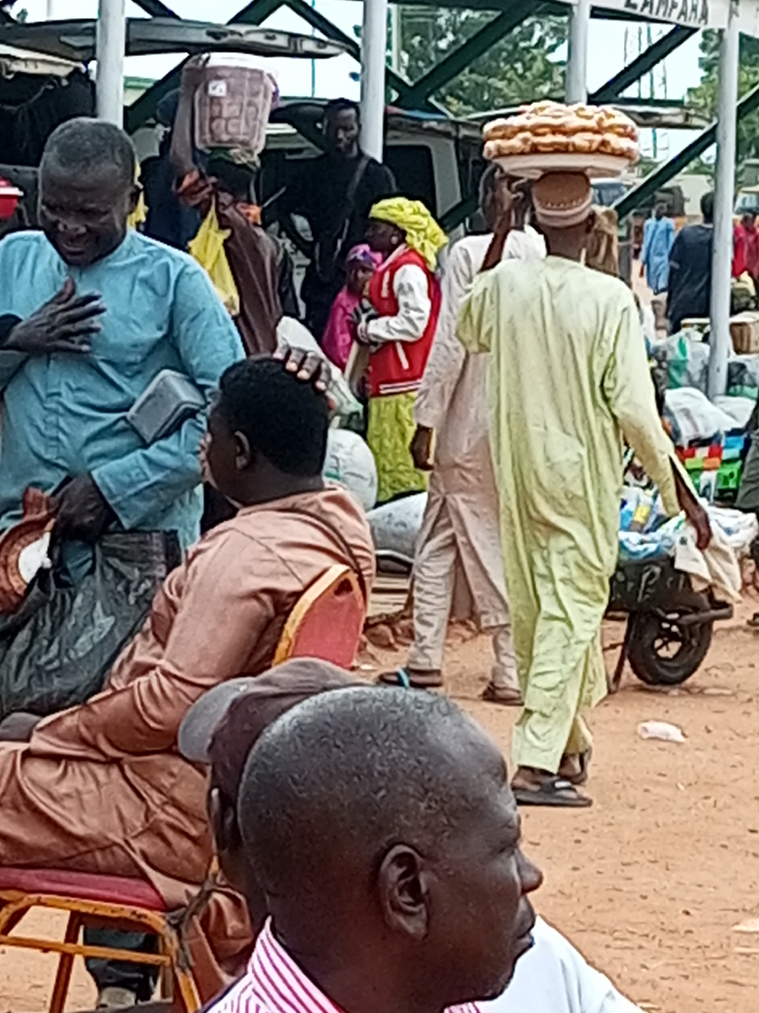 The image depicts a crowded outdoor market scene, likely in the city of Gombe, Nigeria, given the context. In the foreground, a man wearing a light blue garment is shown with his hand on his chest, his expression suggesting distress or a strong emotion. To his right, another man in a brown garment is seated on a chair, looking away from the camera. Further in the background, a man in a pale green garment is carrying a large stack of flatbreads on his head. Other individuals, some partially obscured, are visible amidst stalls and structures typical of a busy market. The ground appears to be dusty earth. The overall atmosphere suggests a lively and potentially chaotic market environment.