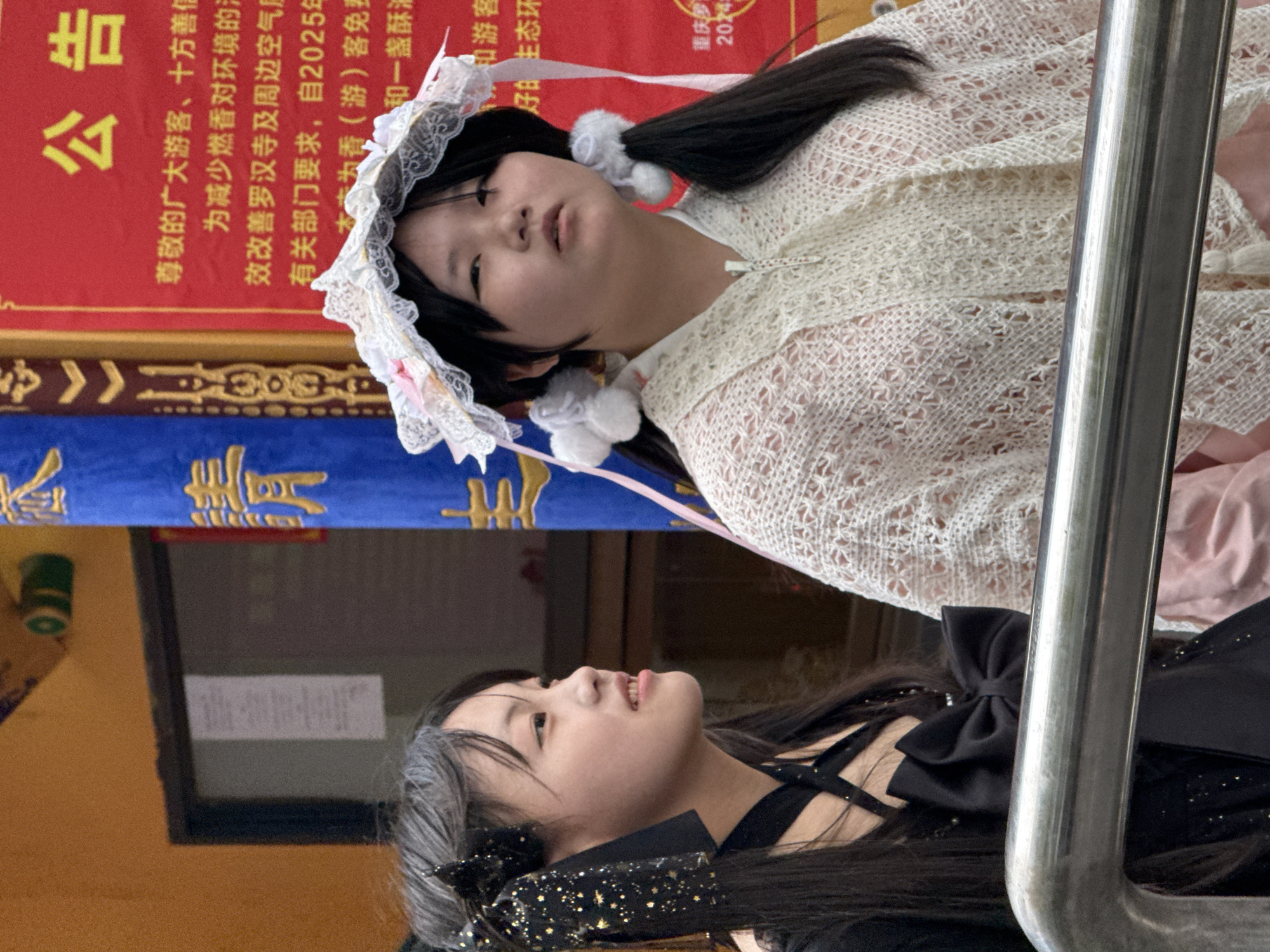 The image shows two young women, one in a black dress with a bow and glittery accents and the other wearing a white, lacy outfit with a matching headpiece and pom-poms. They are outdoors, positioned in front of what appears to be a sign written in Chinese characters. The sign is red with blue trim and gold lettering. The environment includes a brown wall, a metal handrail, and additional text on the sign, suggesting that the scene is likely in a public or religious setting, potentially the Luohan Temple in Yuzhong District, China.