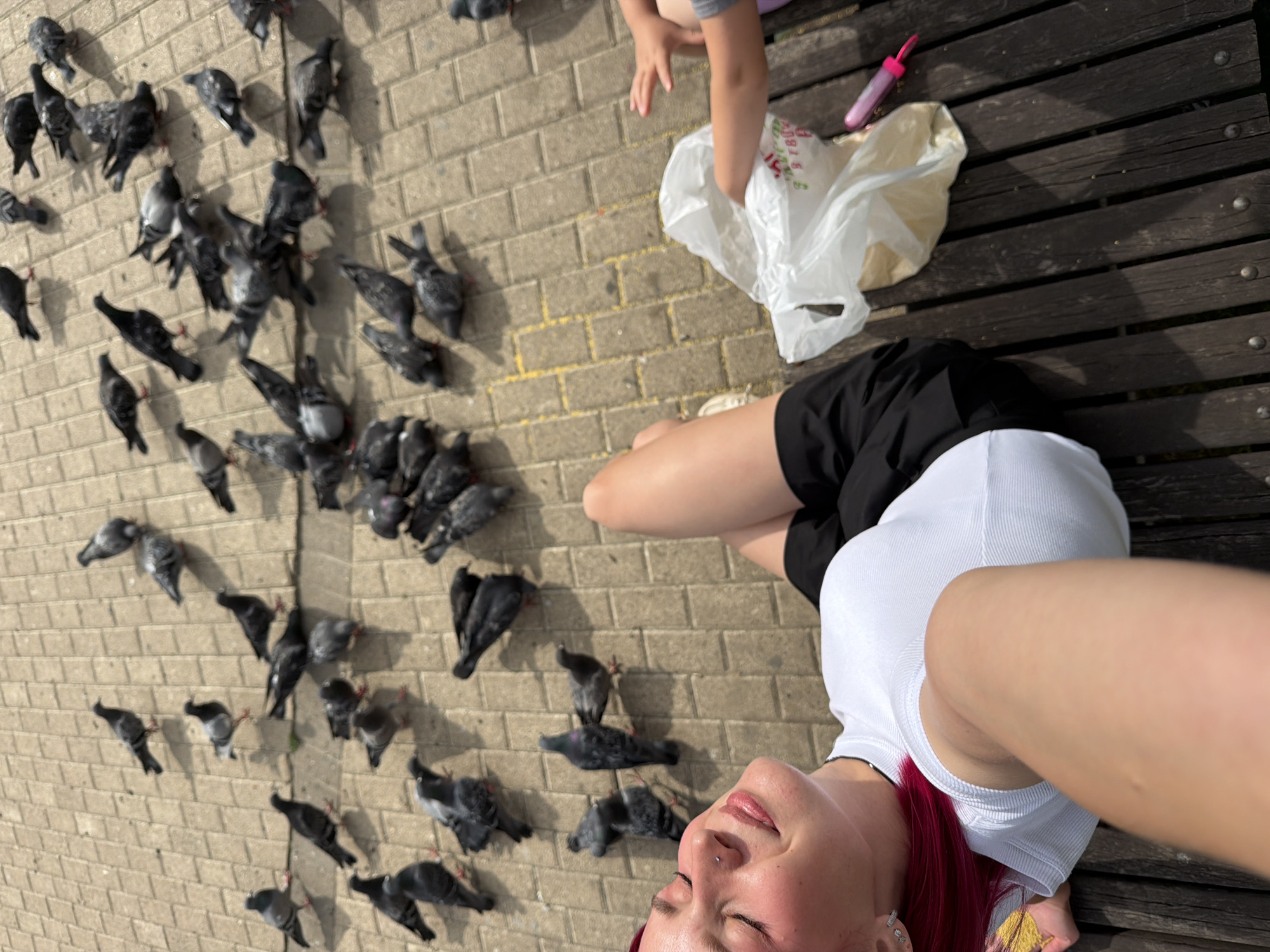 A young woman with bright pink hair and a nose ring is lying on her back on a wooden bench, looking up at the camera. She is wearing a white tank top and black shorts. Her right arm is extended, holding the camera for a selfie. To her left, a child's legs are visible, wearing a grey t-shirt and purple shorts, with their hands reaching towards a white plastic bag on the ground. A pink object, possibly a water bottle, lies on the bench next to the bag. The ground in front of the bench is paved with light brown bricks, and a large flock of pigeons are gathered on the pavement, some pecking at the ground. The scene appears to be outdoors on a sunny day, as indicated by the shadows cast by the pigeons and the bright lighting. The presence of pigeons suggests a public outdoor space, likely a park or plaza. The overall mood is casual and relaxed, with the woman seemingly enjoying the interaction with the birds and the company of the child. No visible text is present in the image.