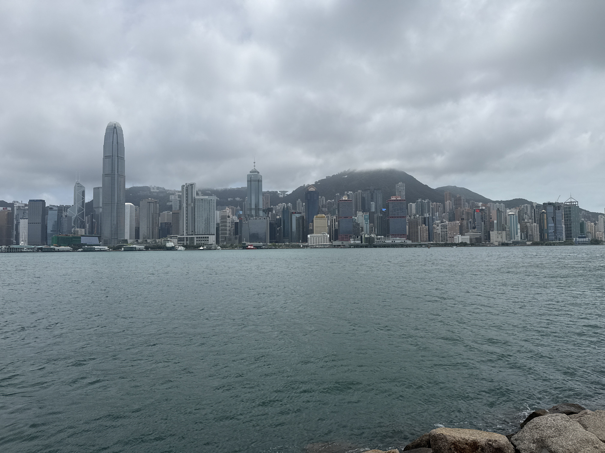The image depicts a cityscape of Hong Kong, China, viewed from across a body of water. The water, which appears to be a bay or harbor, occupies the lower half of the frame and stretches towards the distant skyline. The water is choppy with small waves.
The skyline is dominated by a dense cluster of skyscrapers and high-rise buildings. Several prominent structures stand out, including the International Finance Centre (IFC), recognizable by its tall, slender design. A mountain rises behind the buildings.
The sky is overcast with a thick layer of gray clouds, indicating potentially inclement weather. There are rocks along the shoreline in the lower right. No people are visible in the scene.