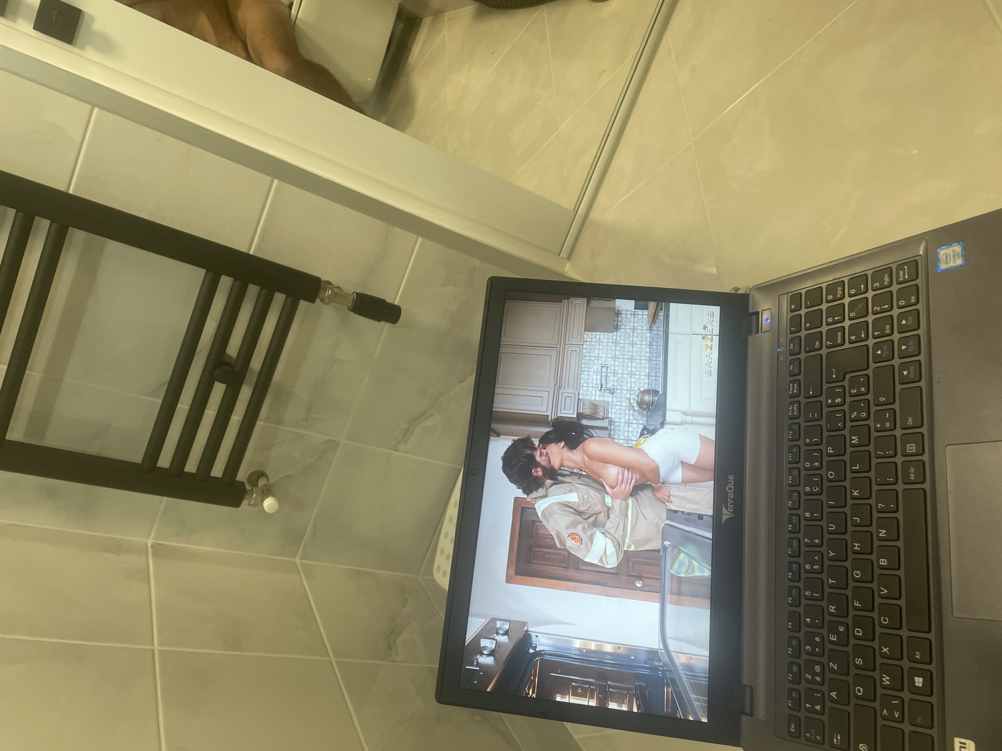 A laptop is placed on a tiled floor in a bathroom. The laptop displays a video of a man in a tan uniform kissing a woman who is topless. In the background of the video, a kitchen scene is visible. The bathroom has light-colored tiles on the walls and floor, and a black heated towel rack is mounted on the wall. A white toilet and a shower door are also partially visible.