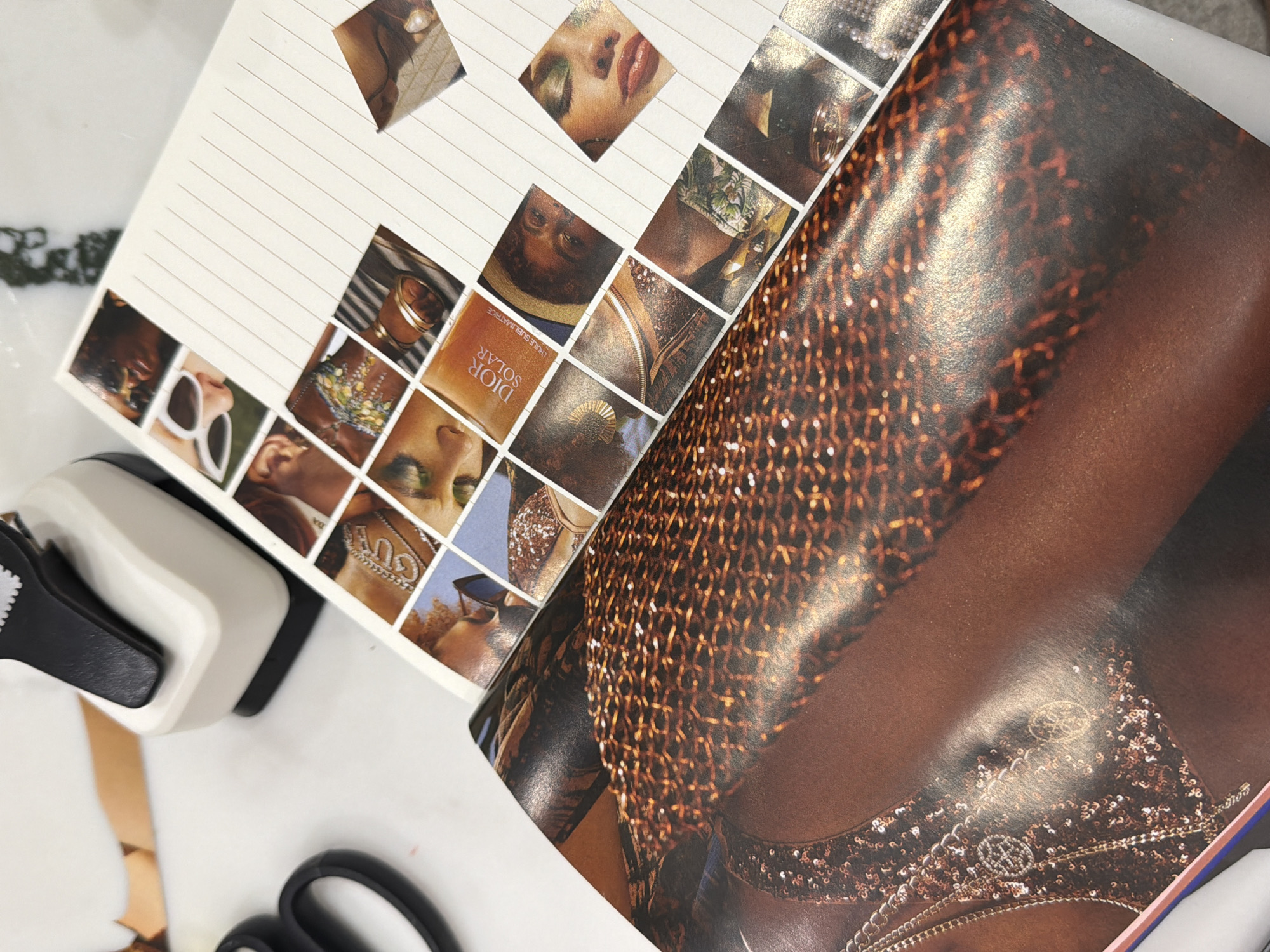 The image shows a close-up of a crafting project in progress. A white notebook with lined pages has a grid of square images glued onto it, depicting fragments of fashion advertisements: close-ups of faces, jewelry, and a Dior product. Overlapping the notebook is a larger magazine page featuring a brown-skinned individual wearing a sparkly, chain-like garment. A white and black hole punch and black scissors are on the left side of the frame, presumably used in the crafting process. Some discarded paper pieces are also visible on the white surface. There is no evidence in the image to indicate this scene is in Yerevan, Armenia.
