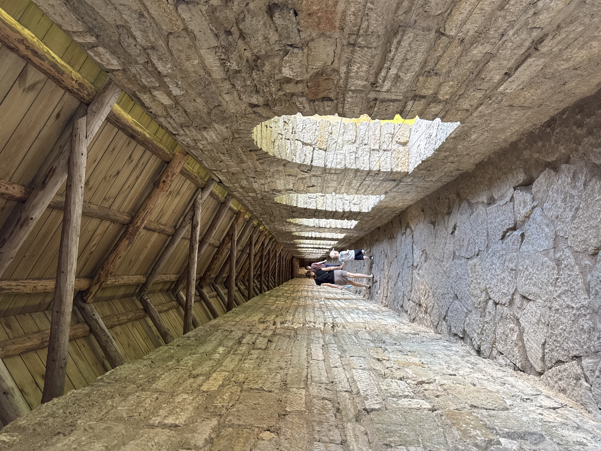 The image shows a person walking through a stone passageway with a wooden roof. The passageway is lined with rough-hewn stone blocks. Arches are visible on the right side, allowing light to enter. The floor is paved with irregularly shaped stones. In the distance, two figures, a woman and a child, are walking away from the camera. The context provided indicates this is a location in the city of Shlisselburg, Russia, specifically within the Oreshek Fortress, a historic site known for its stone walls and fortified structures.