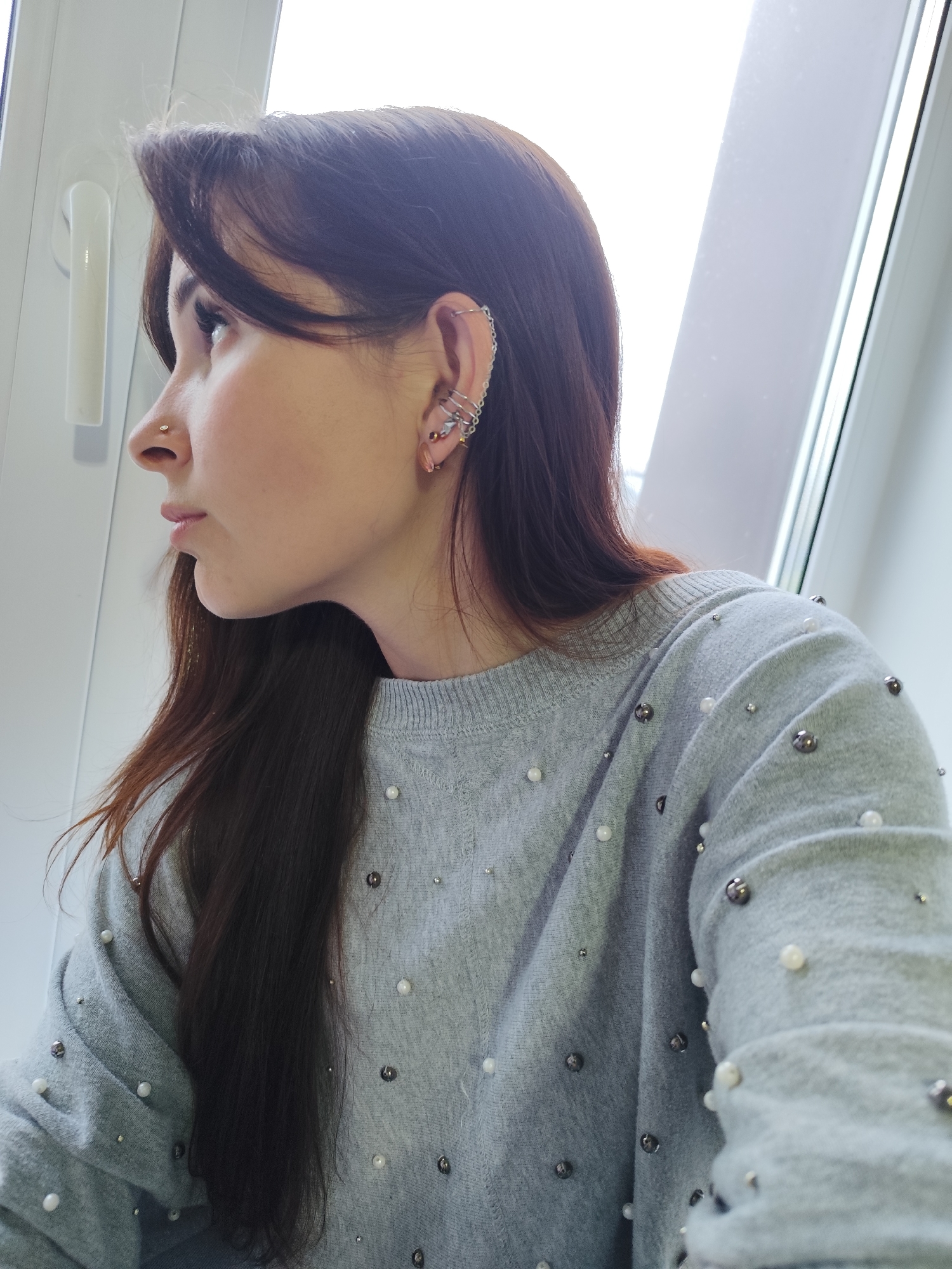 A woman with brown hair and bangs is looking to the left, standing in front of a window. She has a nose piercing and multiple piercings in her right ear, including a cuff that extends along the upper ear. She is wearing a gray sweater decorated with pearls and metal studs. The location is not discernible from the provided image.