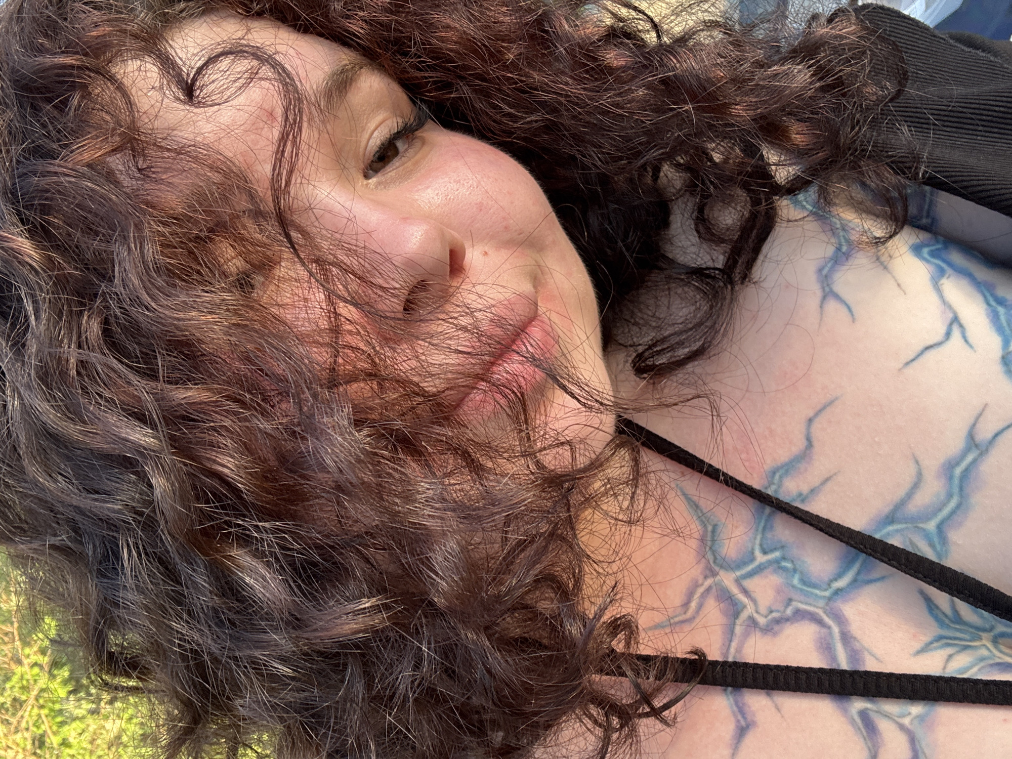 The image is a close-up selfie of a young woman with curly brown hair. She is outdoors, with blurred green foliage in the background. Her face is partially obscured by her hair, but her eyes, nose, and mouth are visible. She is wearing a black garment, possibly a bikini top, and has a tattoo on her chest that appears to be stylized lightning bolts in blue and purple. The lighting suggests it is daytime, and the overall mood is casual and relaxed. There is no specific indication of the location within Surgut, Russia.