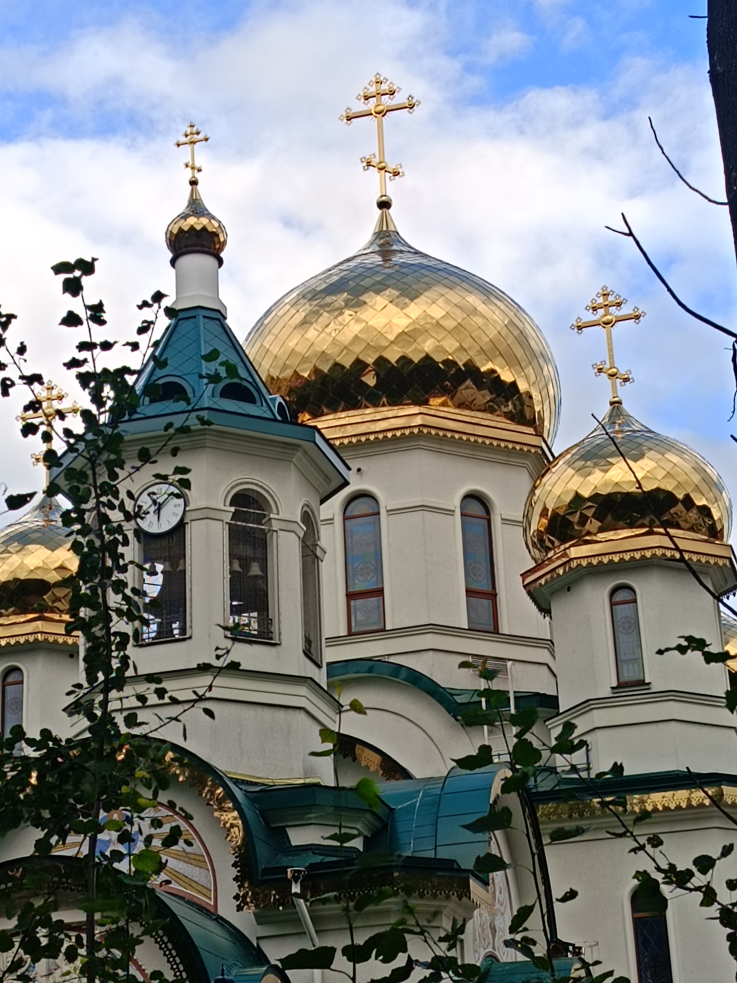 This is a medium-shot, eye-level, outdoor photograph of the Alexander Nevsky Cathedral in Saint Petersburg, Russia, captured on a cloudy day. The cathedral, a prominent landmark, features a distinctive architectural style with multiple golden domes, each topped with a cross.
In the foreground, dark green leafy branches partially obscure the view of the cathedral's white facade. The main subject of the image is the cathedral's structure, showcasing its ornate design. One of the prominent elements is a clock tower with a white clock face and Roman numerals, positioned to the left of the frame. The clock hands indicate the time is approximately 2:45.
The cathedral is adorned with several golden domes of various sizes, reflecting the ambient light. Arched windows with stained glass are visible on the building's walls. The roof sections are a deep teal color, contrasting with the white walls and golden domes. Architectural details such as decorative cornices and trim are visible in gold and white.
The sky is partially cloudy, with patches of blue visible between the white clouds. No people are present in the frame. The overall impression is one of grandeur and religious significance, with the natural elements of the branches adding a touch of organic contrast to the man-made structure.