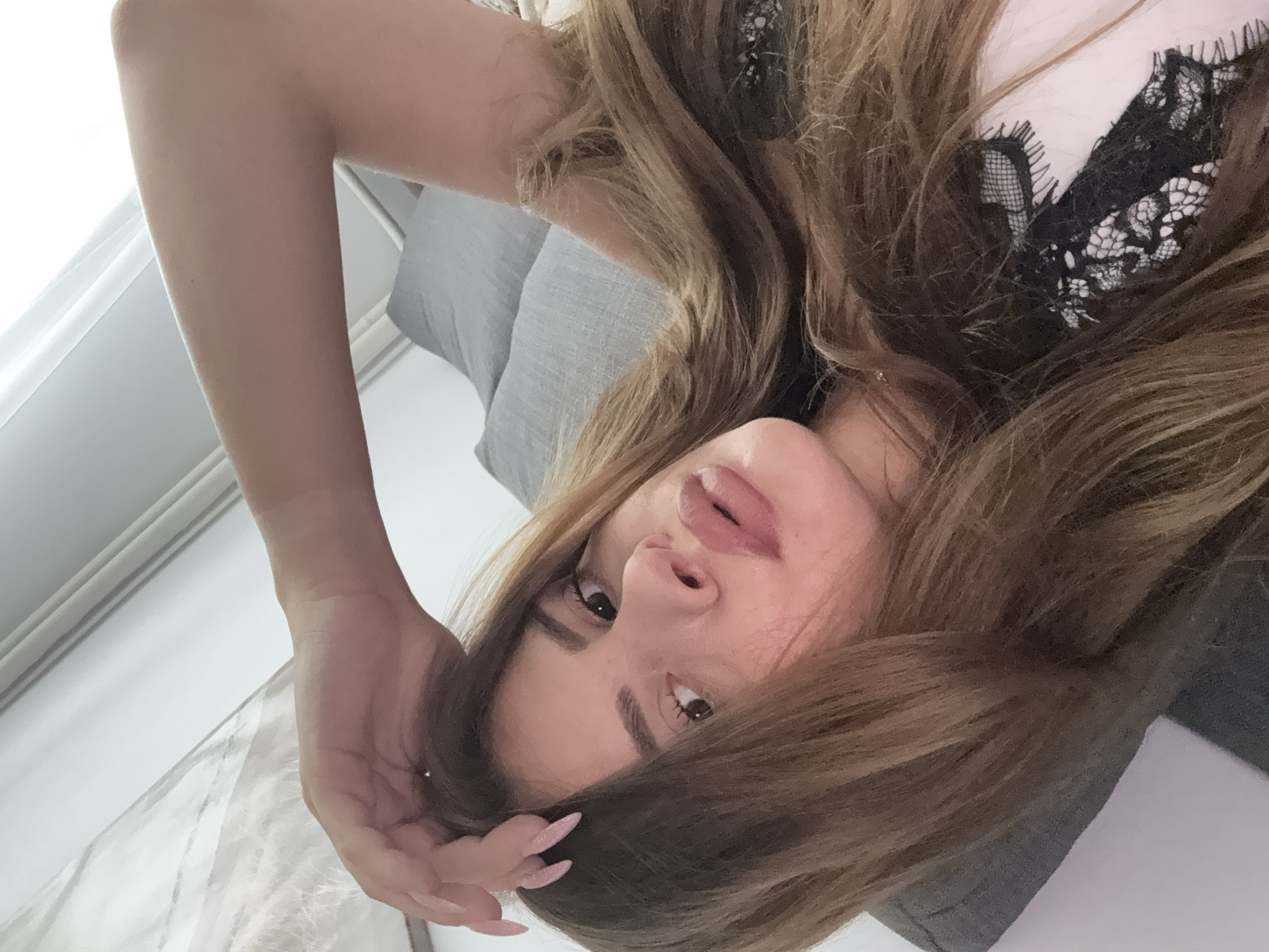 The image shows a close-up, overhead shot of a young woman with long, light brown hair, lying down on a couch. Her right arm is raised and bent, with her hand resting on her head. She is looking at the camera, with her lips slightly parted. She is wearing a light-colored top with black lace detailing. The background appears to be an indoor setting with a grey couch and a white curtain. There is no information in the image to suggest a location in Plovdiv, Bulgaria.