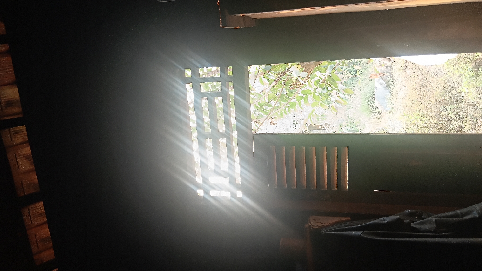 The image depicts the interior of a dark room looking out through a partially open doorway and a barred window. Sunlight streams through the window, creating distinct light rays that illuminate the room and the exterior scene. On the left, a dark garment, possibly a jacket, is hanging on a hook against a wooden surface. To the left of the doorway, a cylindrical object, perhaps a pot or container, is visible. The barred window has a geometric design, allowing glimpses of the outdoor environment, which appears to be a garden or yard with vegetation and a rocky area. The overall impression is of a humble dwelling, possibly in a rural or semi-rural setting within the city of Prabumulih, Indonesia, where natural light plays a significant role in illuminating the interior.