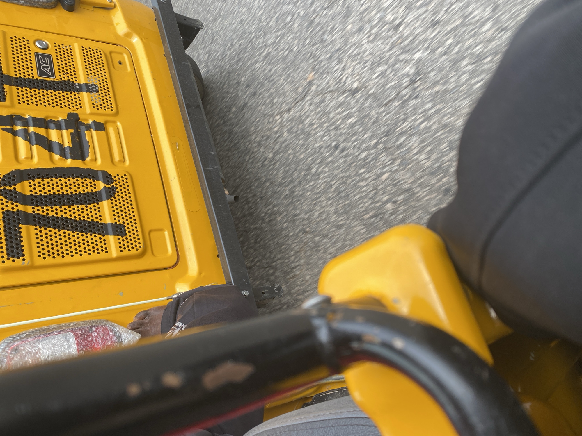 The image shows a close-up view of the rear of a yellow vehicle, possibly a truck or a large utility vehicle, moving on an asphalt road. The vehicle has a perforated metal surface on its back, with large black numbers "701" stenciled across it. The "AC" logo is visible on the left side of the perforated section.
In the foreground, a black metal bar from the vehicle's structure is prominent. To the left and slightly behind this bar, a person's foot is visible, wearing dark trousers. The foot is resting next to an object wrapped in clear bubble wrap, containing what appears to be a red object inside. The person is wearing dark clothing.
The setting is outdoors, with the road surface indicating movement. The lighting suggests it is daytime. The motion blur of the road indicates the vehicle is in transit. No specific weather conditions are discernible. The location context is Jalingo, Nigeria. The overall impression is a snapshot taken from within the vehicle while it is moving.