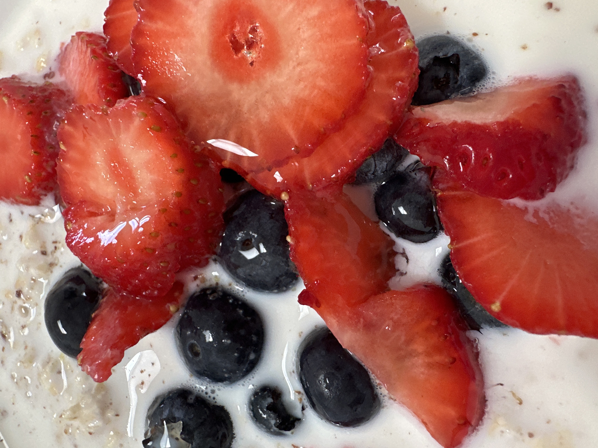 This is a close-up, overhead shot of a bowl of oatmeal topped with sliced strawberries and blueberries. The oatmeal appears to be cooked with milk, creating a creamy base. The berries are fresh and vibrant, with the strawberries sliced into rounds and some larger chunks. The blueberries are whole and dark blue. The lighting is bright, highlighting the textures and colors of the ingredients. There is no indication of the location in Moortown, Ireland or any people present in the image.