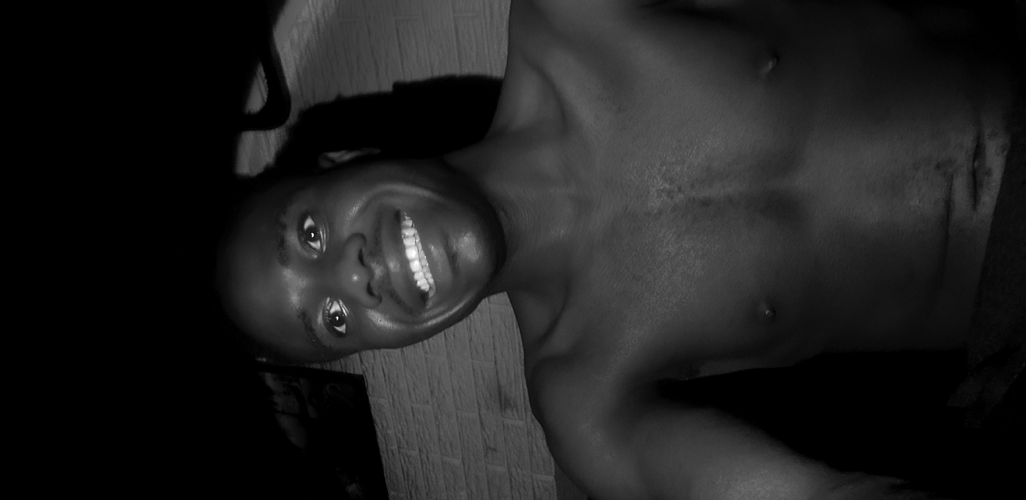 A black and white, close-up selfie shows a shirtless Black man with dark skin, a broad smile, and wide eyes. He is looking up and slightly to the left of the camera. The background includes a textured wall that appears to be brick and a dark, indistinct area above him. The lighting casts shadows on his face and chest, highlighting his features. No specific location in Narayi, Nigeria, can be determined from the image.