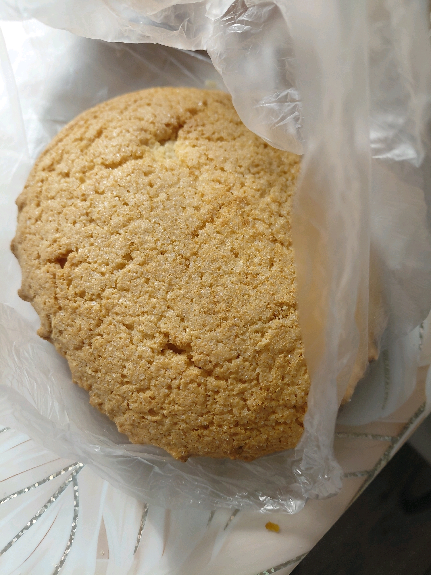 A close-up, overhead shot showcases a single, golden-brown cookie with a textured, granulated sugar surface, nestled within a clear plastic bag. The bag is partially open, allowing the cookie to be prominently displayed. The background is out of focus, but appears to be a table with a patterned surface. The image is well-lit, highlighting the details of the cookie and the transparency of the plastic. The location in Malinovka, Russia, is not discernible from the visual information provided in the image.