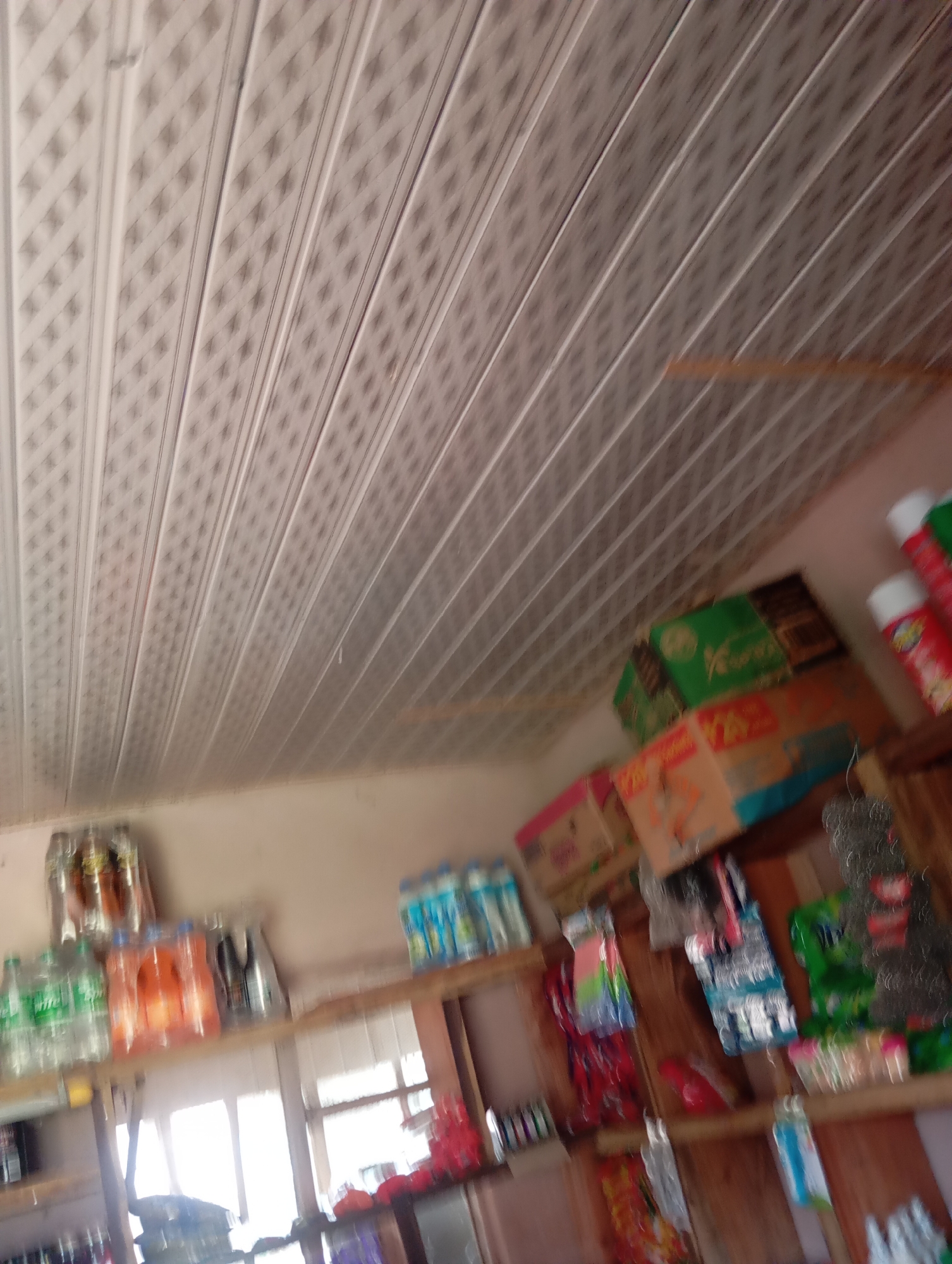 This is a low-angle, slightly blurry, indoor photograph taken in a small shop or convenience store in Jalingo, Nigeria. The ceiling is paneled with a white, textured material, possibly vinyl or PVC, arranged in horizontal strips with a diamond pattern. The walls appear to be a neutral color, possibly beige or off-white.
The shop is stocked with various goods displayed on wooden shelves. On the upper shelves, there are several bottles and packs of beverages, including what appears to be Sprite and orange-flavored drinks. There are also some boxed items, one of which is green and has some text on it. Further to the right, there are aerosol cans, one red and white, and another red with yellow accents.
On lower shelves, more packaged goods are visible, including what look like bags of snacks or other small items. In the foreground, towards the bottom right, there are some cleaning supplies or brushes with gray bristles. The lighting in the store suggests it might be daytime, with light coming from windows in the background, which are partially obscured by shelves. The overall impression is of a modestly stocked local shop.