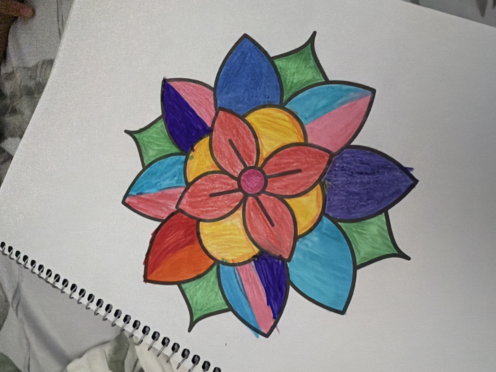 The image shows a drawing of a colorful flower on a white spiral-bound notebook. The flower is centrally located and features multiple layers of petals in various vibrant colors including red, orange, yellow, green, blue, pink, and purple. The petals are outlined in black. The notebook appears to be on a surface that is out of focus, possibly a bed or a blanket. The background of the image is also out of focus, showing some textured fabric. There are no people visible in the image, and no specific location in a city is depicted.