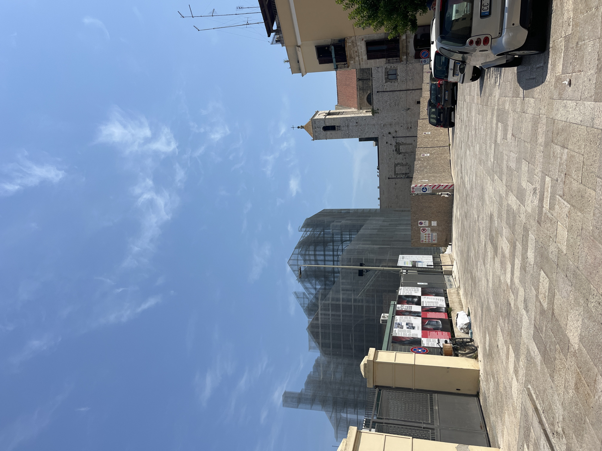 The image depicts a street scene in Bari, Italy, under a clear blue sky with wispy clouds. In the foreground, a paved area with large, light-colored stones is visible. To the left, a beige building facade with a metal gate stands adjacent to a lamppost. Behind the gate, a bicycle with a basket is leaning against a fence. A series of posters are displayed on the fence, some with images and text, including one that reads "SAN PIETRO DELLE FOSSE."
Dominating the background is a large structure covered in scaffolding and netting, suggesting ongoing construction or renovation. To the right of the scaffolding, a stone church tower with a cross on top rises against the sky, its architecture characteristic of historical Italian buildings. Adjacent to the church, a cream-colored building with balconies is visible. Several cars are parked on the street, including a white Smart car in the foreground. The environment appears to be an urban setting, likely a public square or street in the historic district of Bari.