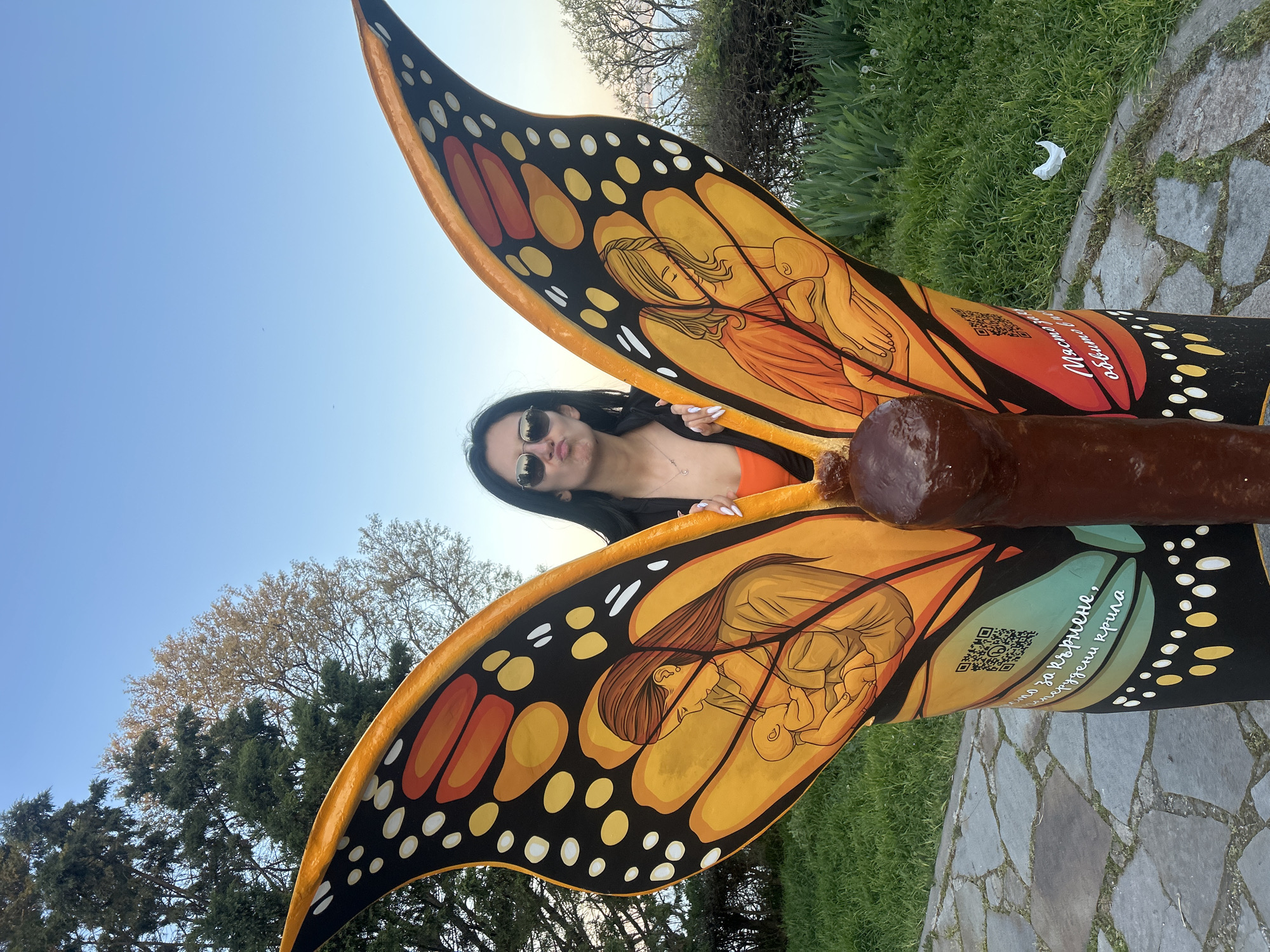 A person is standing behind a butterfly wing sculpture in a park-like setting. The person is wearing sunglasses and an orange top. The sculpture features monarch butterfly wing designs, including drawings of a woman holding a baby. One wing includes a QR code and writing in Bulgarian. The ground is covered in gray stone pavers, with grass and trees visible in the background. Based on the writing and context, this image is likely taken in Burgas, Bulgaria, which is a city known for such public art installations.