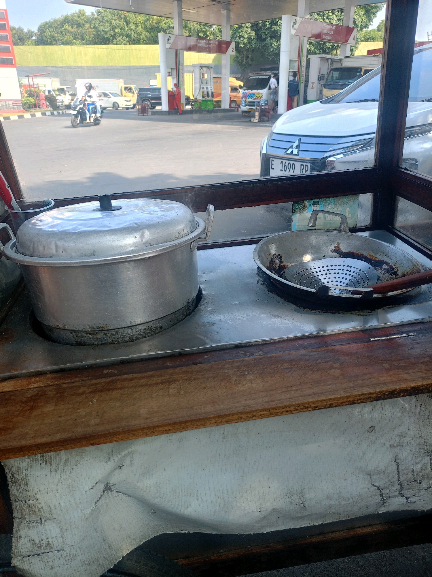 The image captures a close-up view of cooking equipment on a food cart, set against the backdrop of a busy gas station. In the foreground, a large, aluminum pot with a lid rests on a stovetop. To its right, a well-used frying pan with a slotted metal strainer inside is visible. The cart itself appears to be made of wood and metal, with a stained and worn fabric lining underneath.
In the background, a gas station with red and white signage is operational, with cars and motorcycles filling up. A white car with the license plate "E 1699 RP" is parked near a pump. Several other vehicles, including vans and a motorcycle with riders, are also present at the station. People can be seen interacting with the pumps and walking around. The environment is characterized by bright daylight, indicating an outdoor setting. The presence of a food cart in a gas station suggests a potential vendor operating near a high-traffic area in Cirebon, Indonesia.