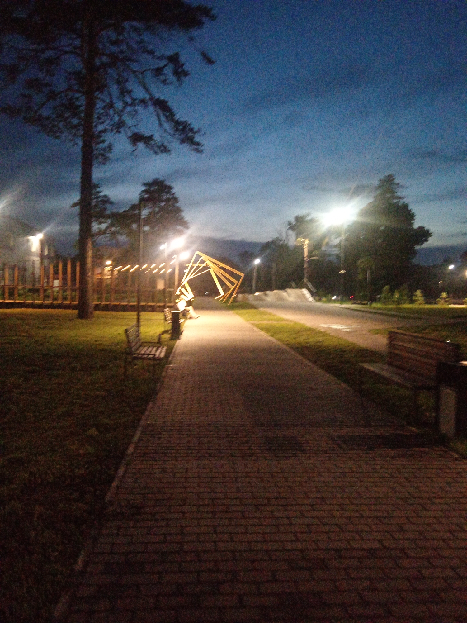 An outdoor scene at dusk features a paved walkway lined with grass and trees. Streetlights illuminate the path and a wooden sculpture. Benches are placed along the walkway. A wooden fence with vertical planks runs along the left side of the scene. The sky is dark blue, indicating the transition from day to night.