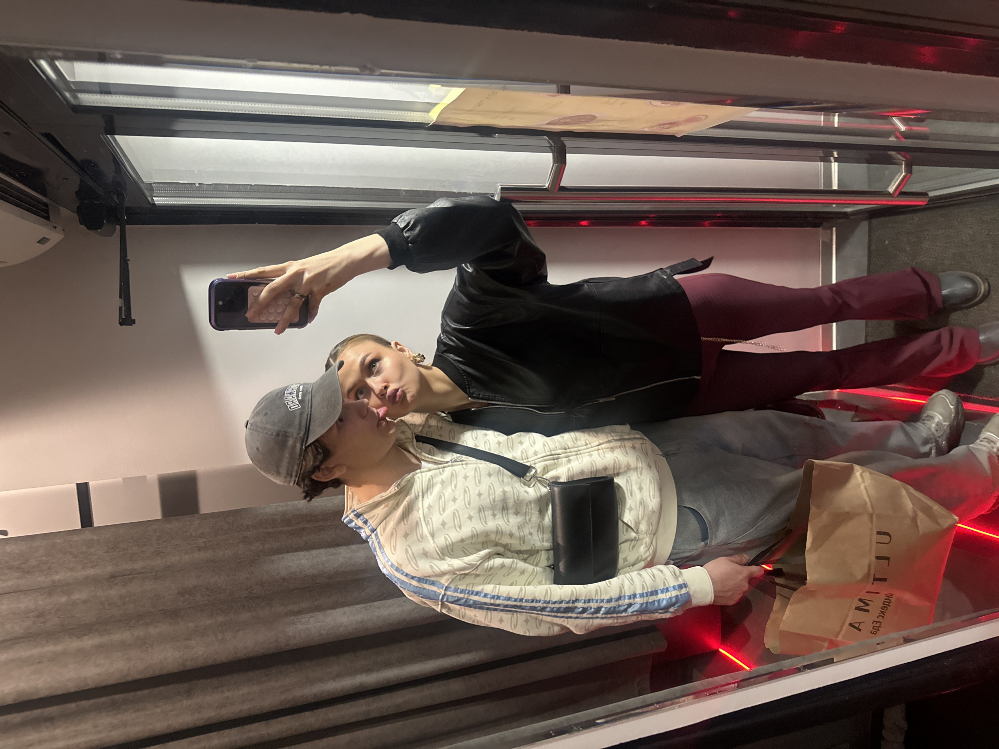 Two people are taking a selfie in an elevator. The person on the left is wearing a grey baseball cap, a white and blue patterned zip-up hoodie, light blue jeans, and silver sneakers, with a black bag slung across their chest. The person on the right is wearing a black leather jacket, maroon pants, and grey shoes, holding a black smartphone to take the photo. They are positioned in what appears to be a glass elevator, based on the visible floor, and there is a brown paper bag with "AMITIU" printed on it in mirrored text, suggesting the location is Krasnoyarsk, Russia, as "AMITIU" is a brand in that city. Red LED lights illuminate the floor. There's a grey cushioned surface along the back wall of the elevator. Above, there is a metal railing and a fluorescent light panel. A partially visible paper is attached to the ceiling.