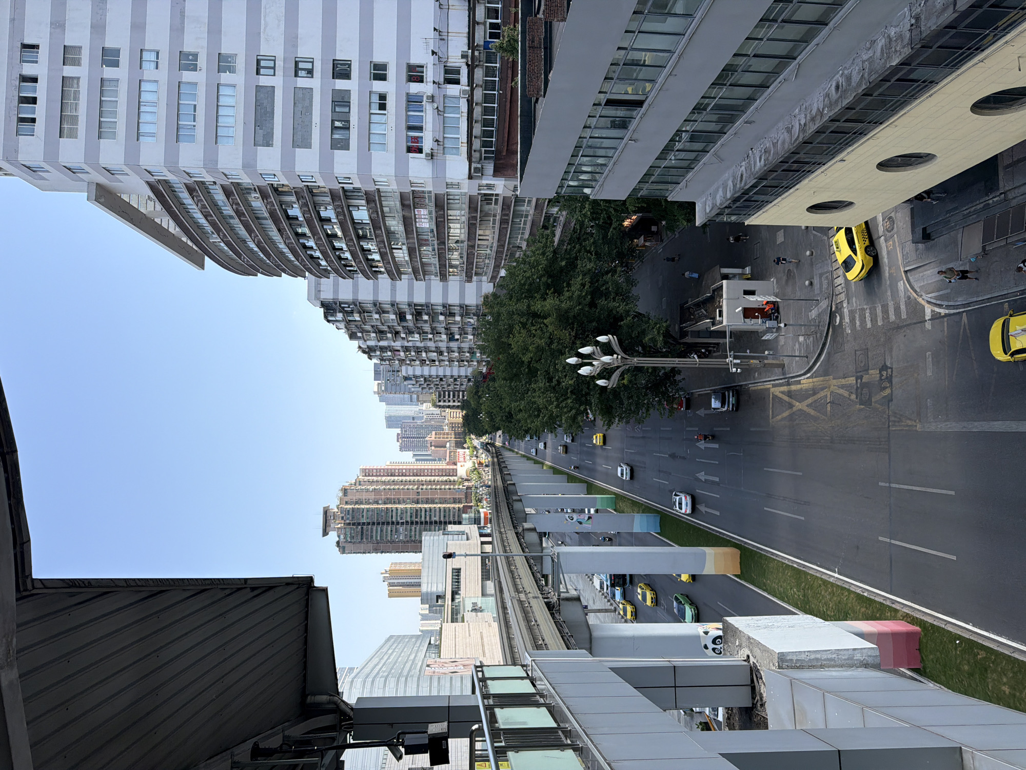 This is a high-angle, eye-level shot of a street in Jiulongpo District, China, on a clear day. The scene is dominated by a raised monorail track and supporting pillars that run parallel to a multi-lane road. Several vehicles, including yellow taxis and other cars, are visible on the road, some of which appear to be in motion. Tall, modern buildings, some with distinctive striped facades and balconies, line the right side of the street. Lush green trees are interspersed with the urban structures, particularly along the side of the road. On the left side of the image, part of a structure with a sloped, corrugated roof and a railing is visible, likely a station or elevated walkway. Pedestrians are sparsely scattered on the sidewalks and crossing the street. A street lamp with multiple bulbs stands near the road. The overall impression is of a busy urban environment with elevated transportation infrastructure.