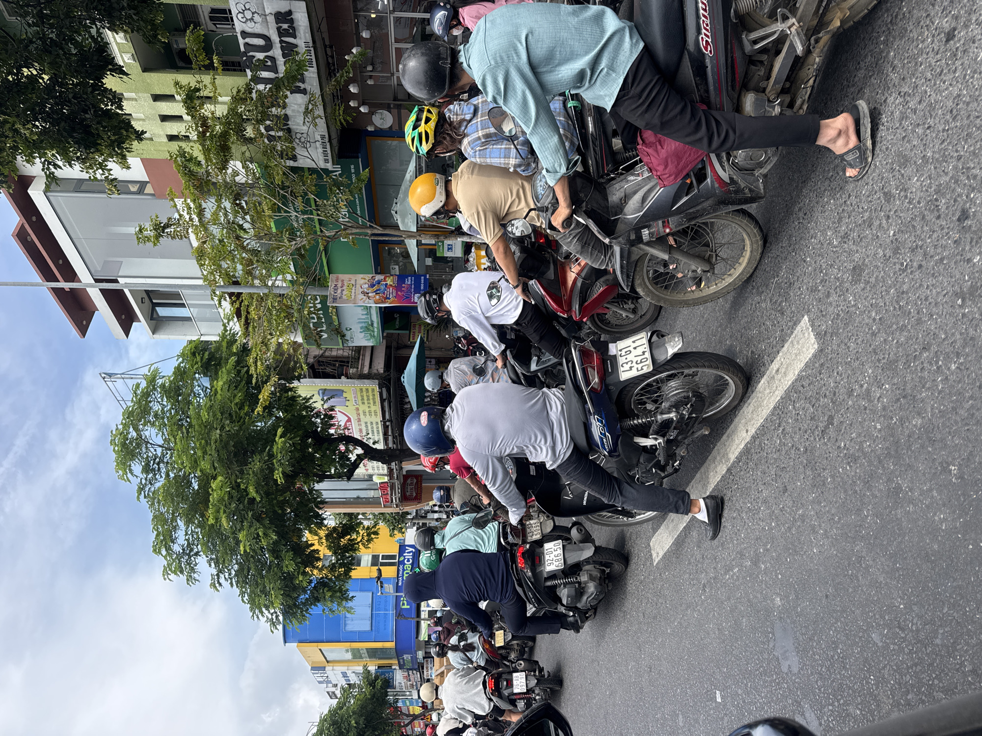 The image shows a line of motorbikes and their riders at a standstill on a street in Đà Nẵng, Vietnam. The riders are a mix of men and women, wearing casual clothing and helmets. The motorbikes are various makes and models, tightly packed together. Street signage and buildings are visible in the background, including storefronts and apartments. The street appears to be moderately busy with traffic. The license plates on the motorbikes are visible, indicating local registration.