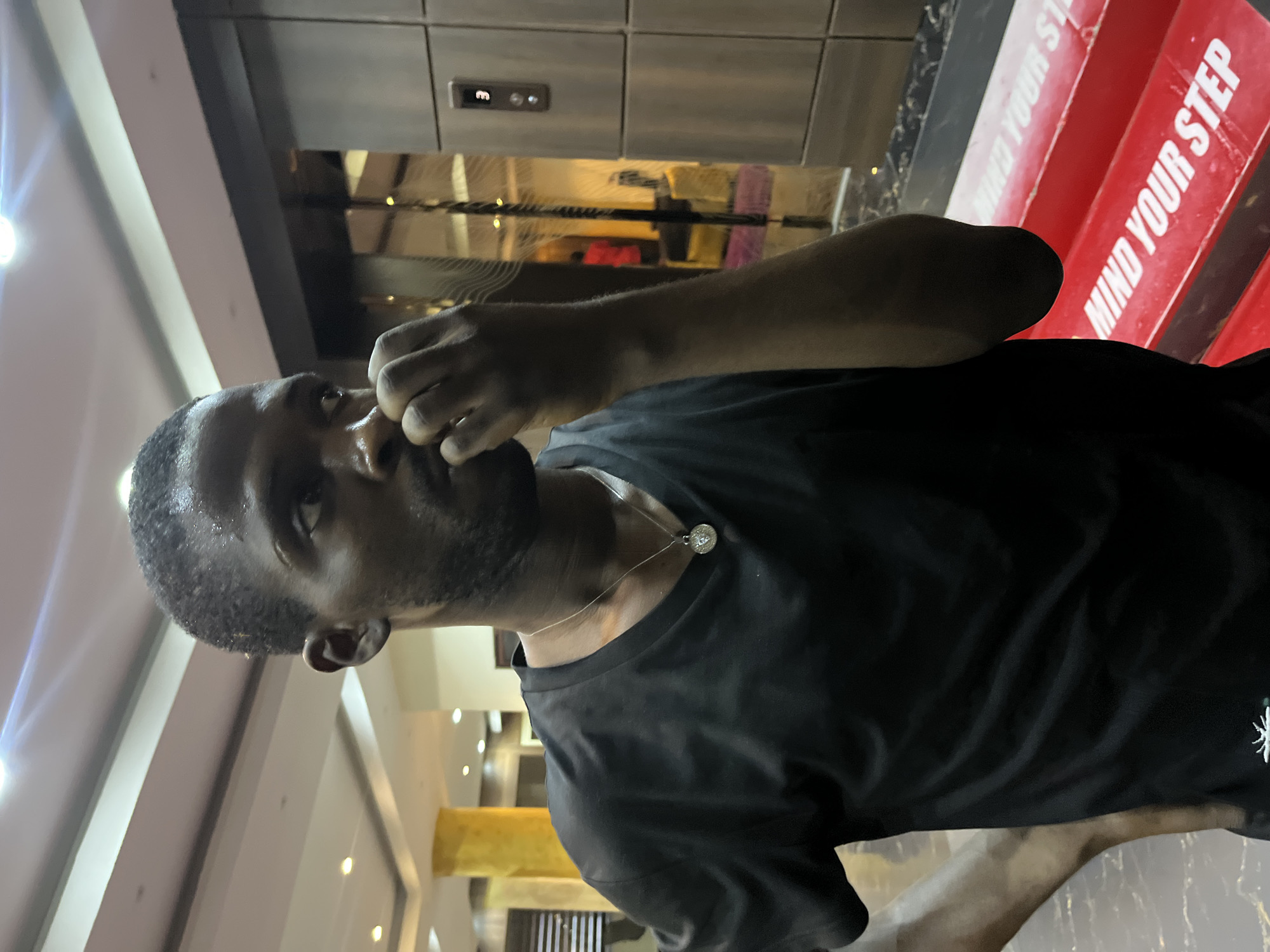 In Abuja, Nigeria, a young Black man with short, dark hair and a light beard is photographed indoors. He is wearing a black t-shirt with a silver pendant necklace. His right hand is raised to his face, with his fingers touching his lips. He appears to be looking to his right, with sweat visible on his forehead. In the background, a metallic elevator door is visible, with the number "3" illuminated. The man is standing on a patterned floor. Red, rectangular objects with the white text "MIND YOUR STEP" are visible in the lower right portion of the image. The environment appears to be a well-lit indoor space with modern architecture.