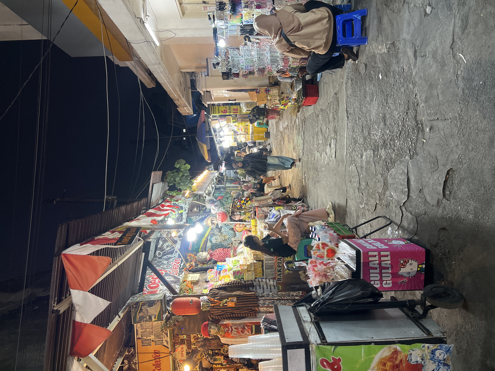 The image depicts a street market at night in Yogyakarta, Indonesia. The narrow alleyway is lined with stalls displaying various goods, including clothing, accessories, and food items. Red and white pennant flags are strung across some of the stalls, possibly indicating a local celebration or event. The ground is paved with cracked concrete, and overhead, a tangle of electrical wires crisscrosses the dark sky. Several people are visible, some browsing the stalls and others sitting or standing. One person, wearing a brown hijab and beige hoodie, is seated on a blue plastic stool, facing away from the camera. Another individual in a hijab and patterned jacket is walking down the alley. The stalls are illuminated by artificial lights, creating a vibrant and bustling atmosphere. A cart with a pink cooler featuring Hello Kitty imagery is prominently displayed in the foreground.
